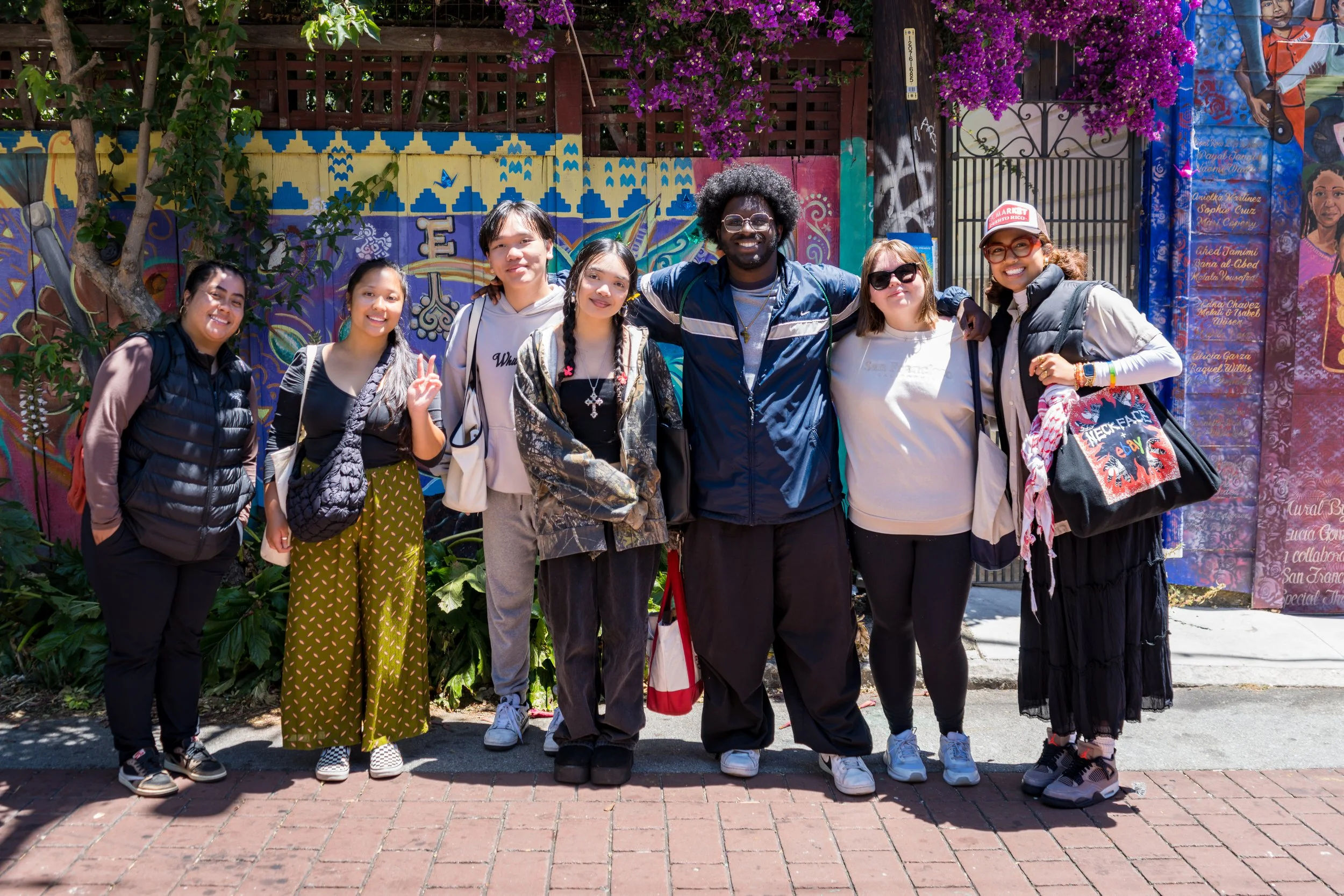 Group of eight diverse people standing outdoors in front of a colorful mural, smiling for a photo.