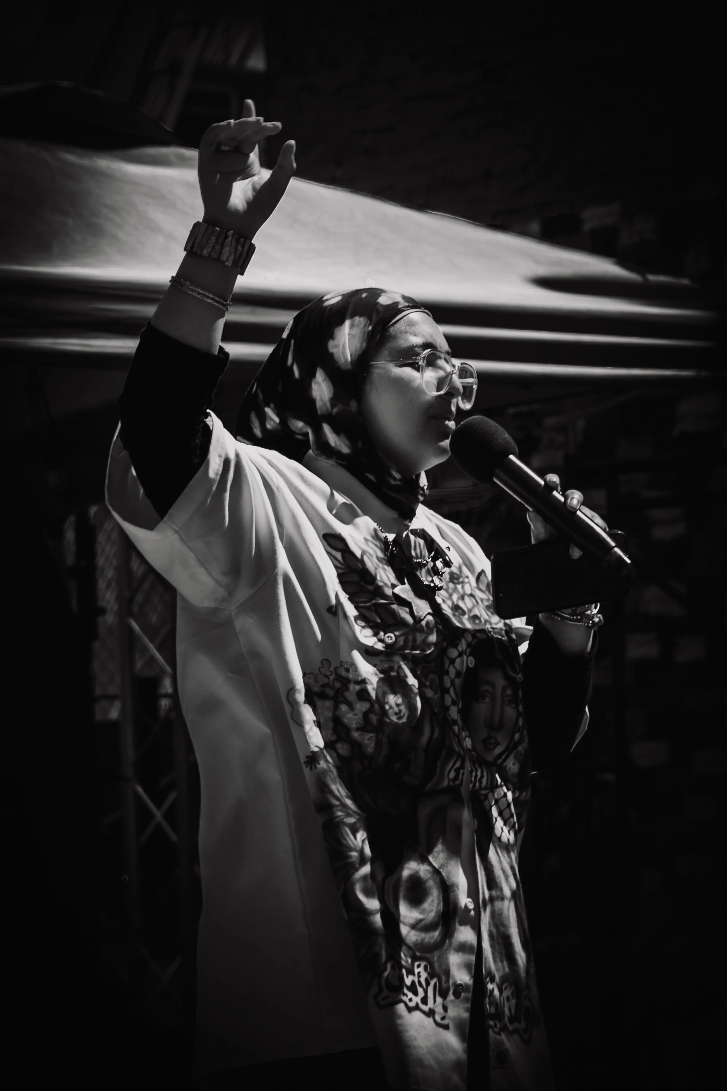 A woman wearing glasses and a patterned headscarf stands in front of a microphone, raising her right hand, speaking or singing. The image is in black and white.