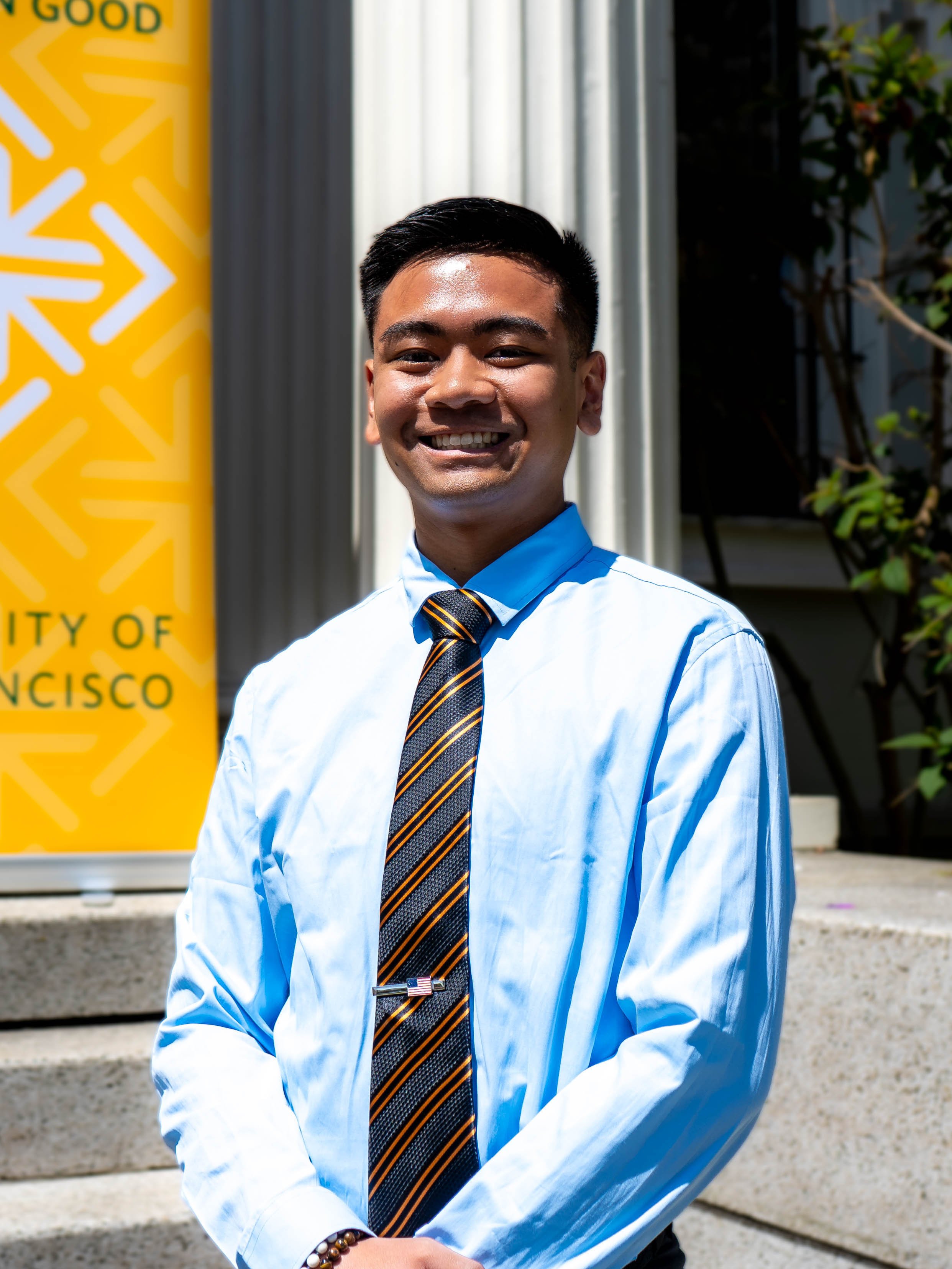 A young man in business attire standing outdoors, smiling, with a yellow sign indicating the City of San Francisco in the background.