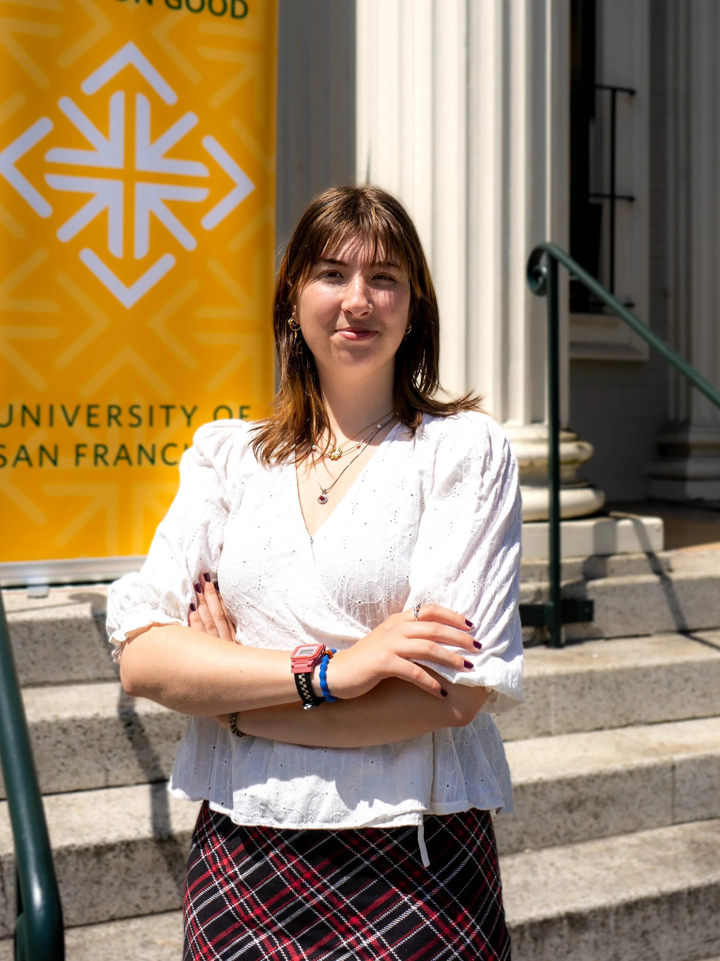 A young woman with shoulder-length brown hair standing in front of a yellow banner with a white snowflake logo and the words 'University of San Francisco.' She is wearing a white blouse, plaid skirt, and accessories.