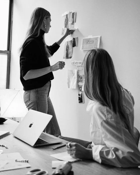 Two women in an office, one standing and pinning papers on a wall, and the other sitting at a desk with a laptop and papers, working on a project.