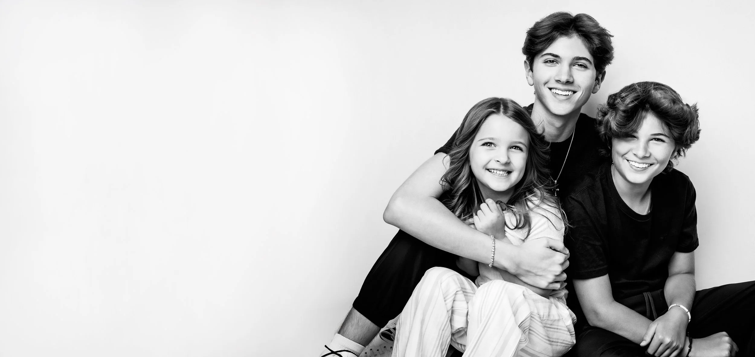 Black and white photo of three young people smiling and sitting close together, with a plain background. The person in the middle is a young woman with long hair, and to her right is a young girl with curly hair and to her left is a young man with wavy hair.
