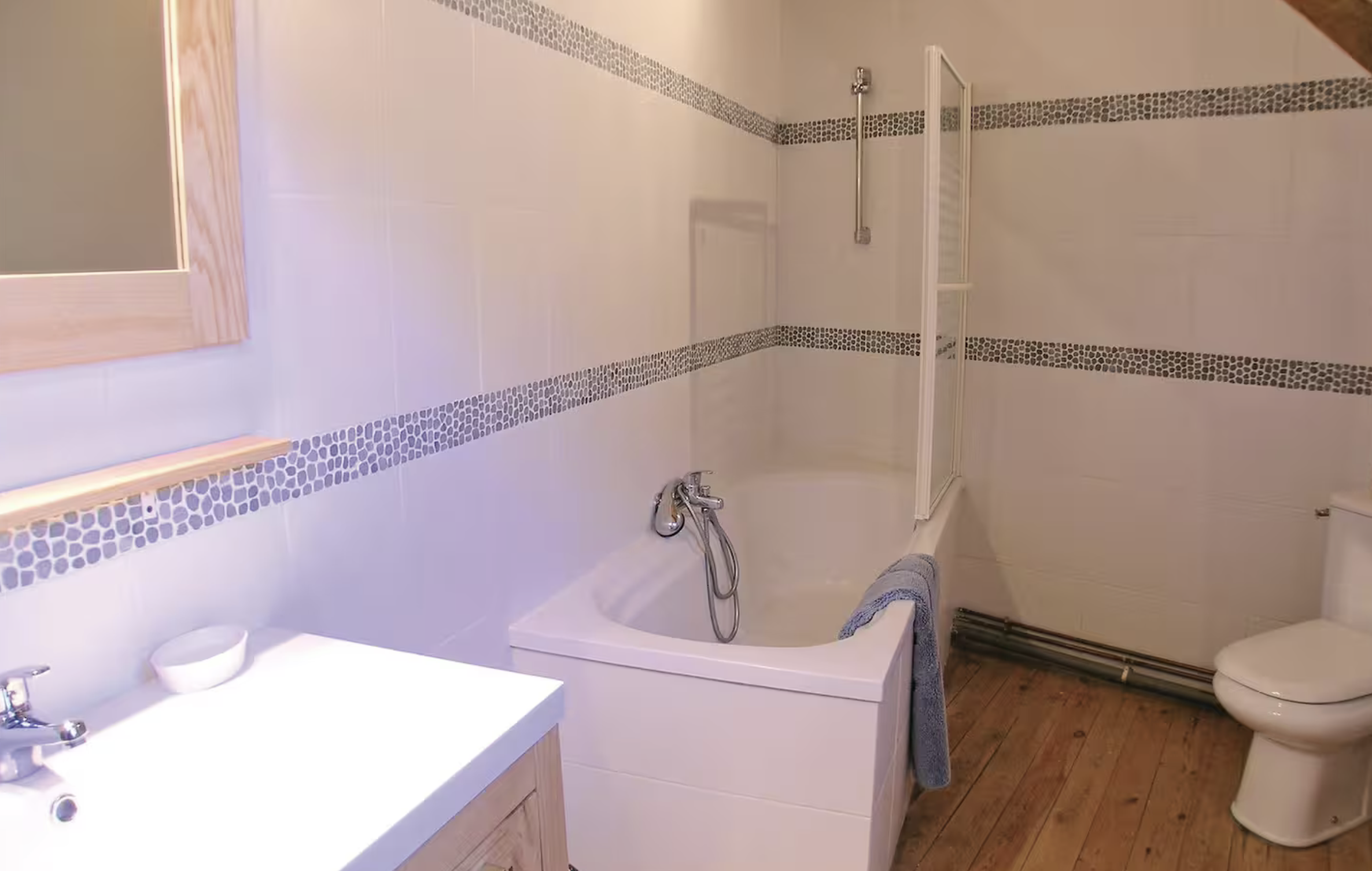 Salle de bain avec baignoire, lavabo avec miroir, toilettes et sol en bois, murs carrelés avec bordure de petits mosaïques.