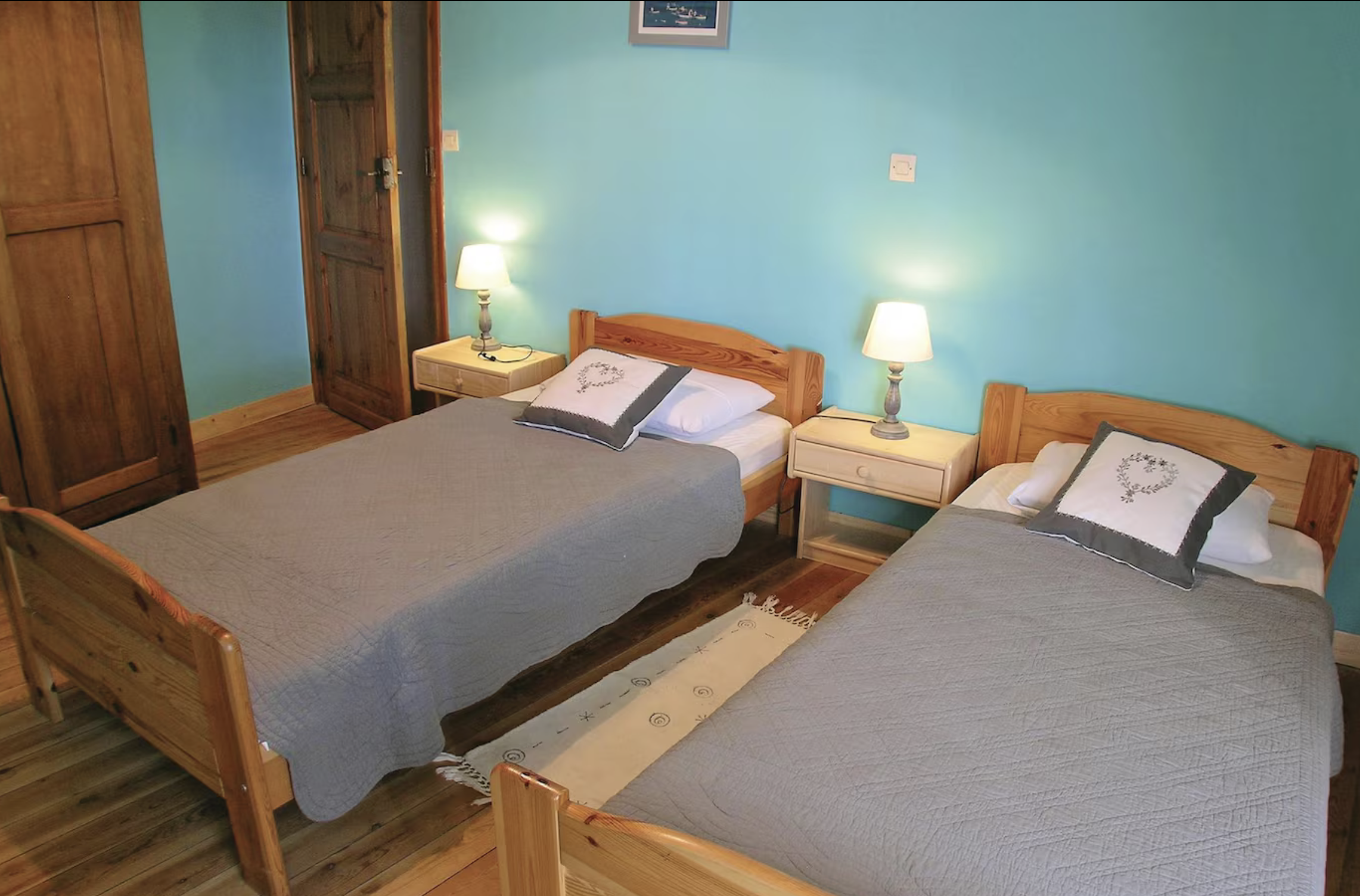 Chambre avec deux lits en bois, couvertures gris clair, coussins avec motif de cœur, lampes de nuit, meubles en bois, mur turquoise, sol en bois.
