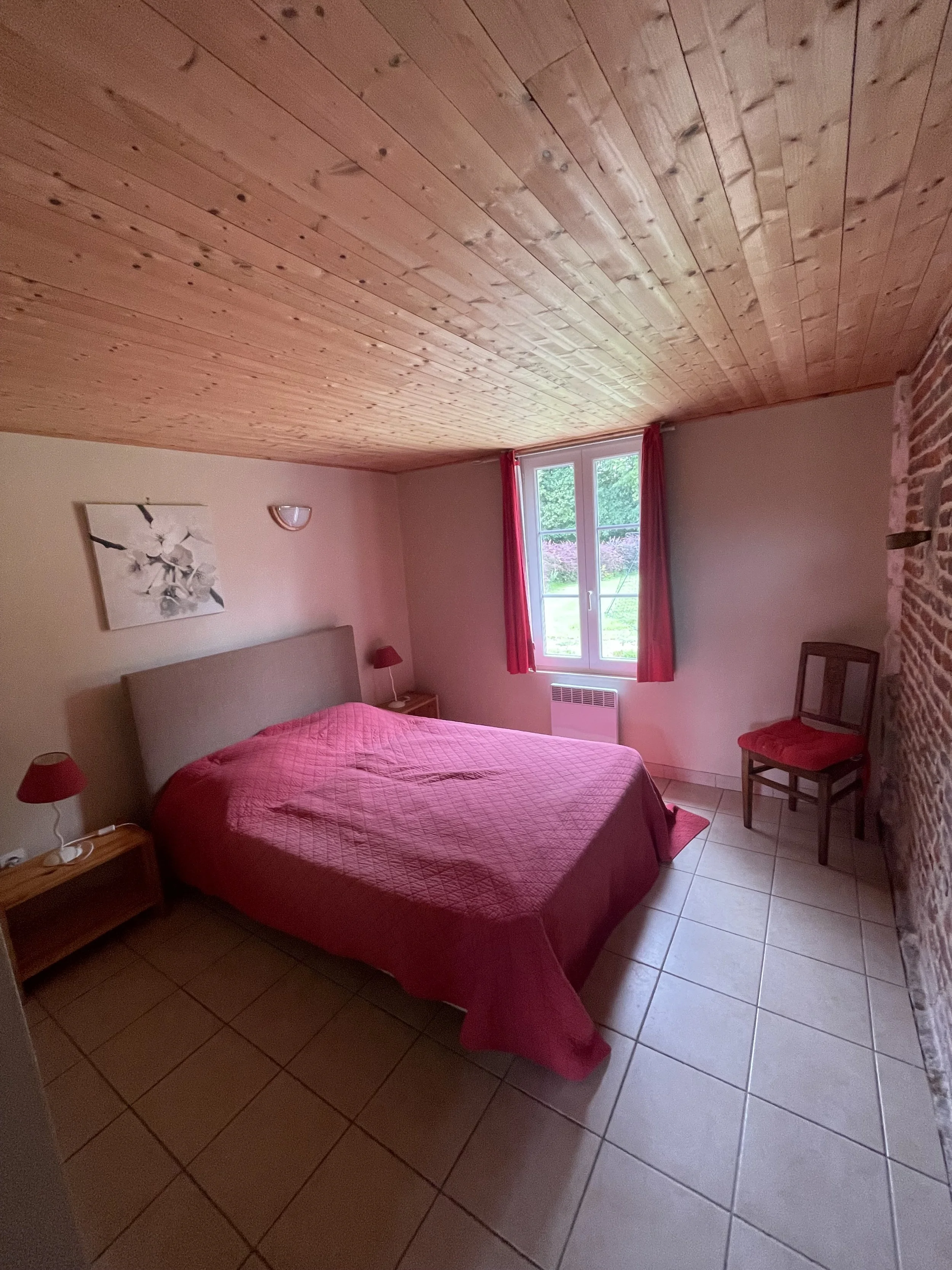Chambre avec un lit avec une couverture rose, deux petites tables de chevet avec lampes roses, une fenêtre avec rideaux roses, un tableau avec des fleurs et un mur en briques. Le plafond en bois et le sol en carrelage sont visibles.