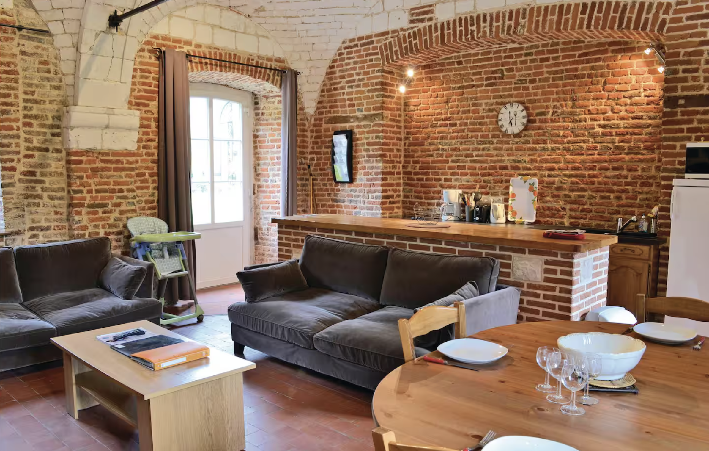 Salon avec murs en briques apparentes, canapés en cuir, table à manger en bois avec assiettes et verres, cuisine partiellement visible en arrière-plan.