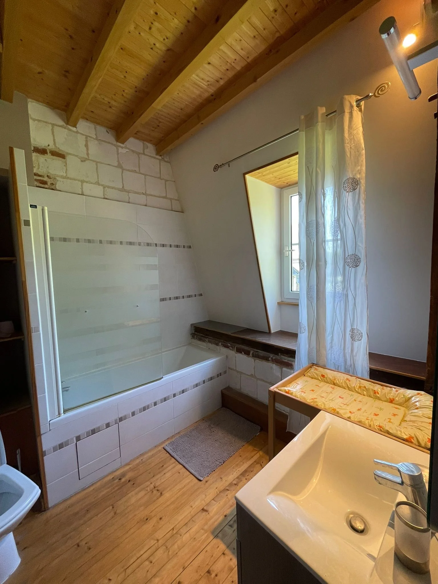 Salle de bain avec baignoire, fenêtre inclinée, rideau blanc à motifs, lavabo, tapis au sol et mur en pierre et bois.