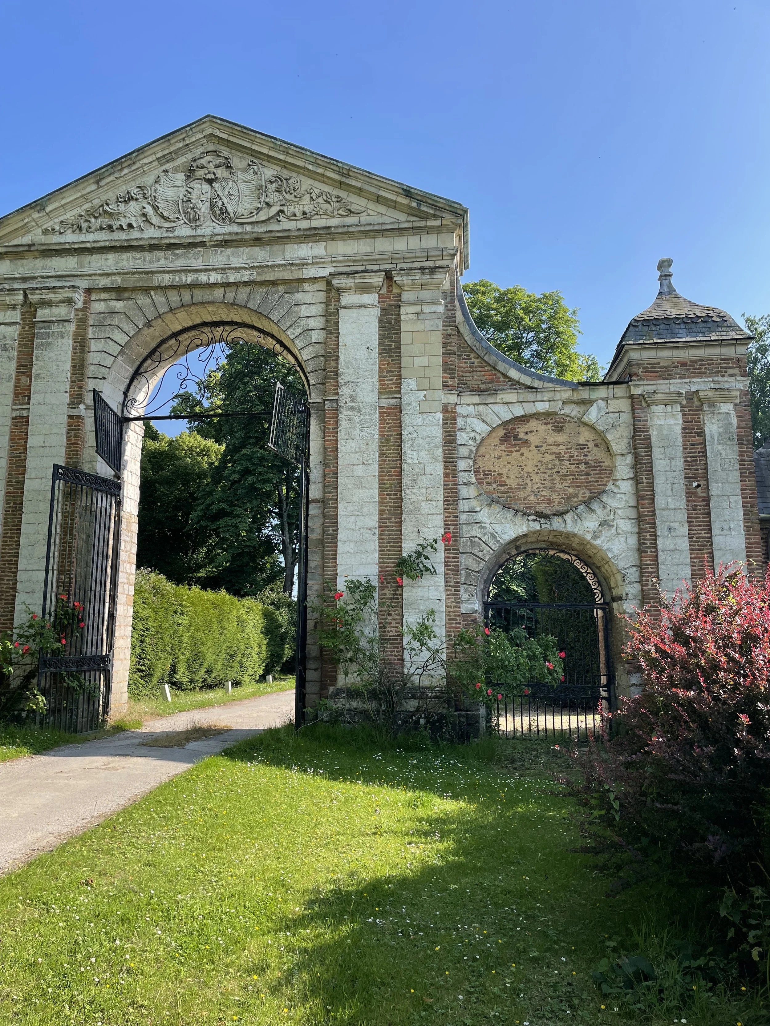 Porte en pierre ancienne avec deux portails en fer forgé, entourée de végétation et de fleurs, sous un ciel bleu clair.
