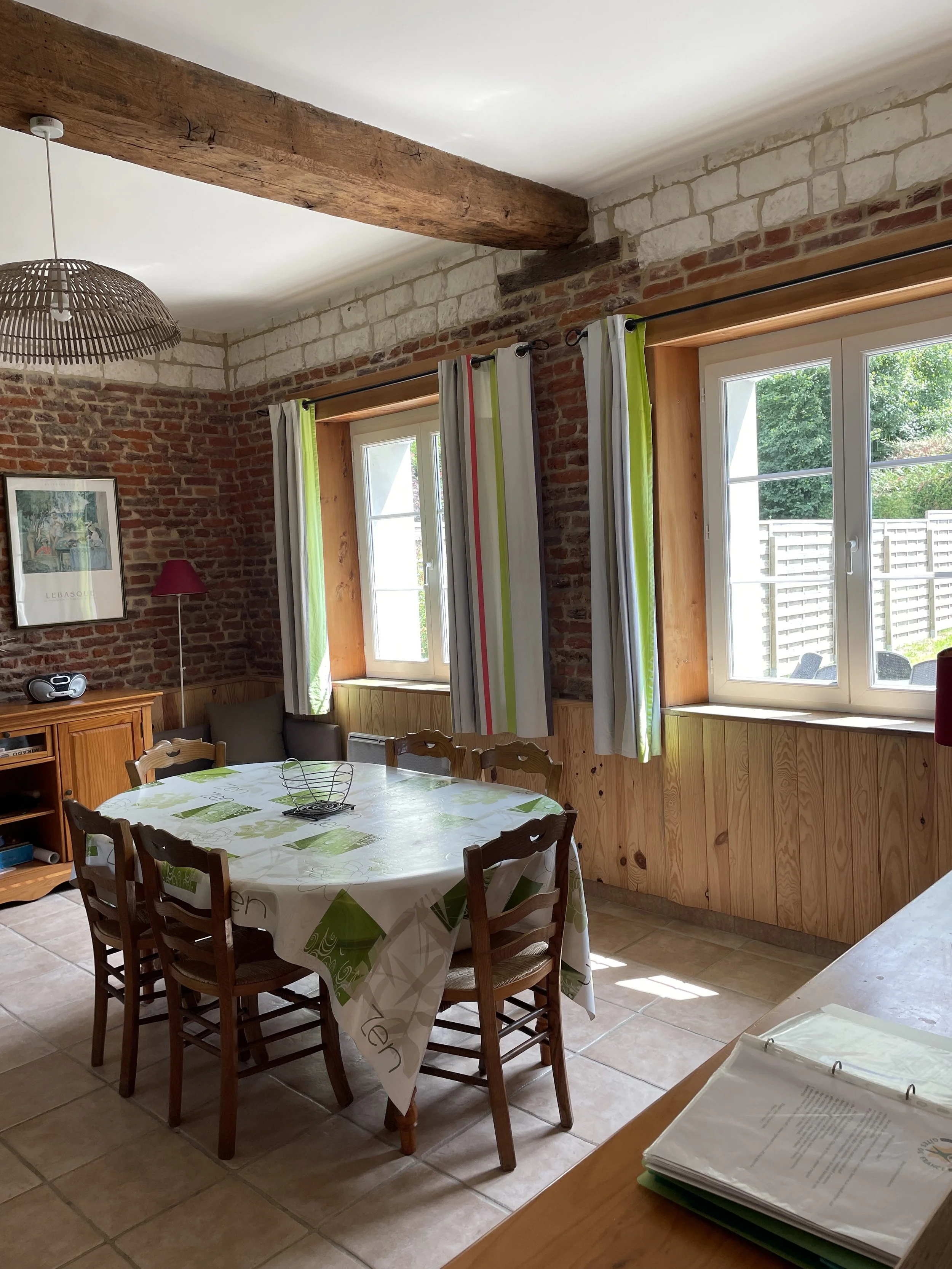 Salle à manger avec table ovale, chaises en bois, murs en briques, fenêtres avec rideaux verts et blancs, plafond avec poutres en bois, arti de décoration et un meuble en bois