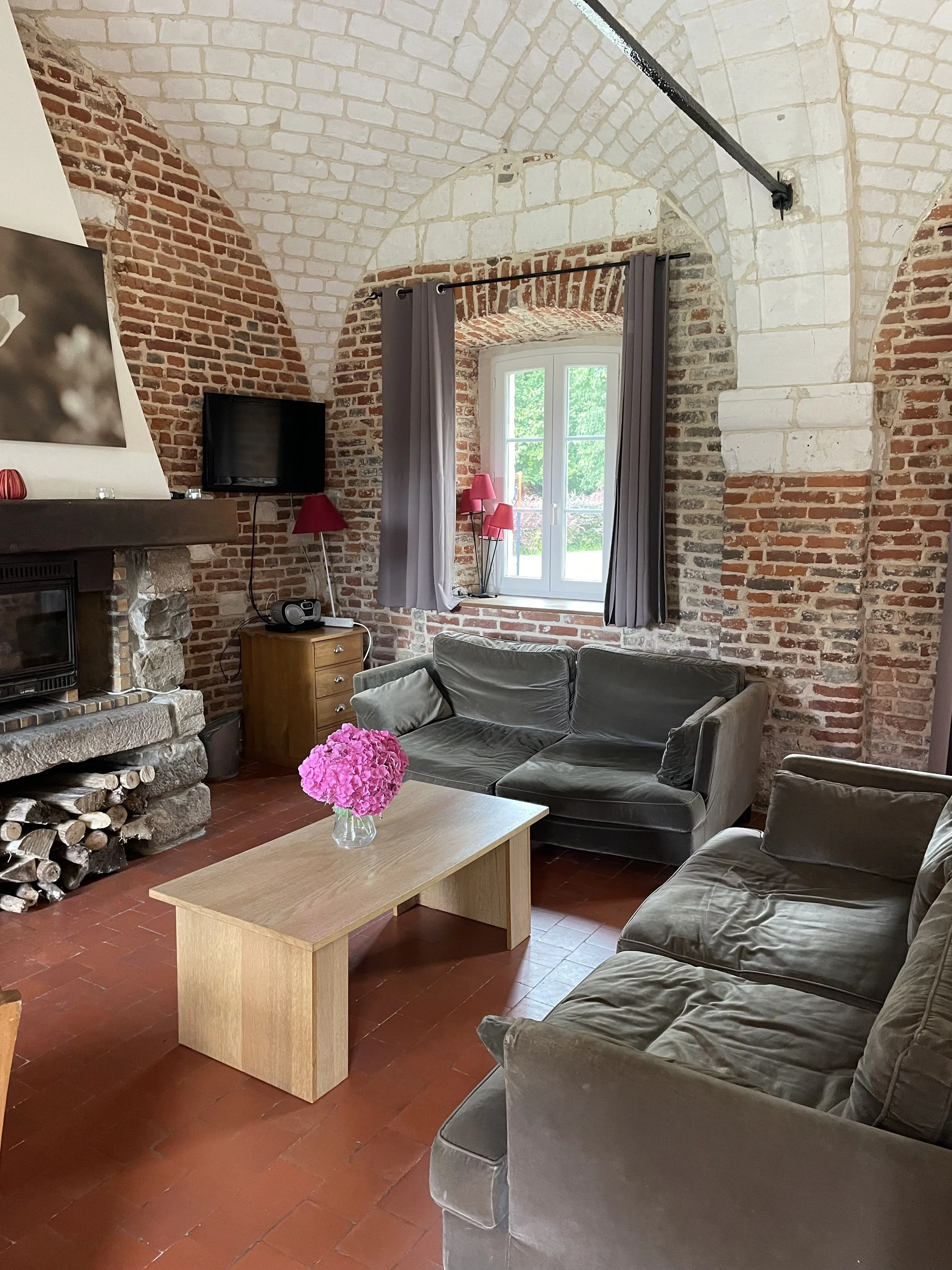 Salon avec canapés gris, cheminée en pierre, table en bois avec vase de fleurs roses, murs en briques rouges et blanches, fenêtre avec rideaux gris et lampes rouges.