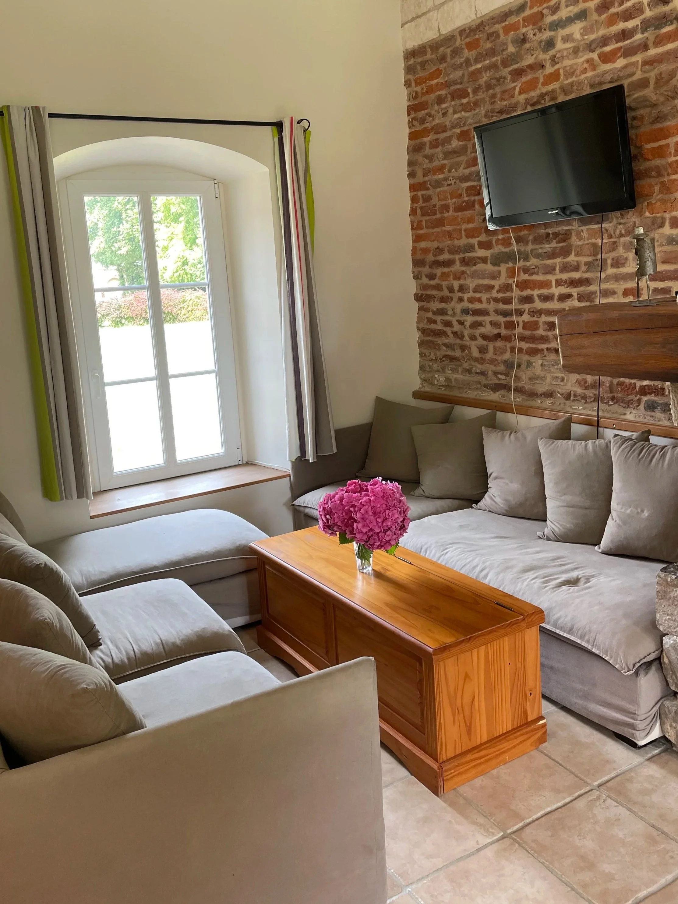 Salon avec canapé gris, table en bois avec bouquet de fleurs roses, fenêtre avec volets, mur en briques, télévision accroché au mur en briques.