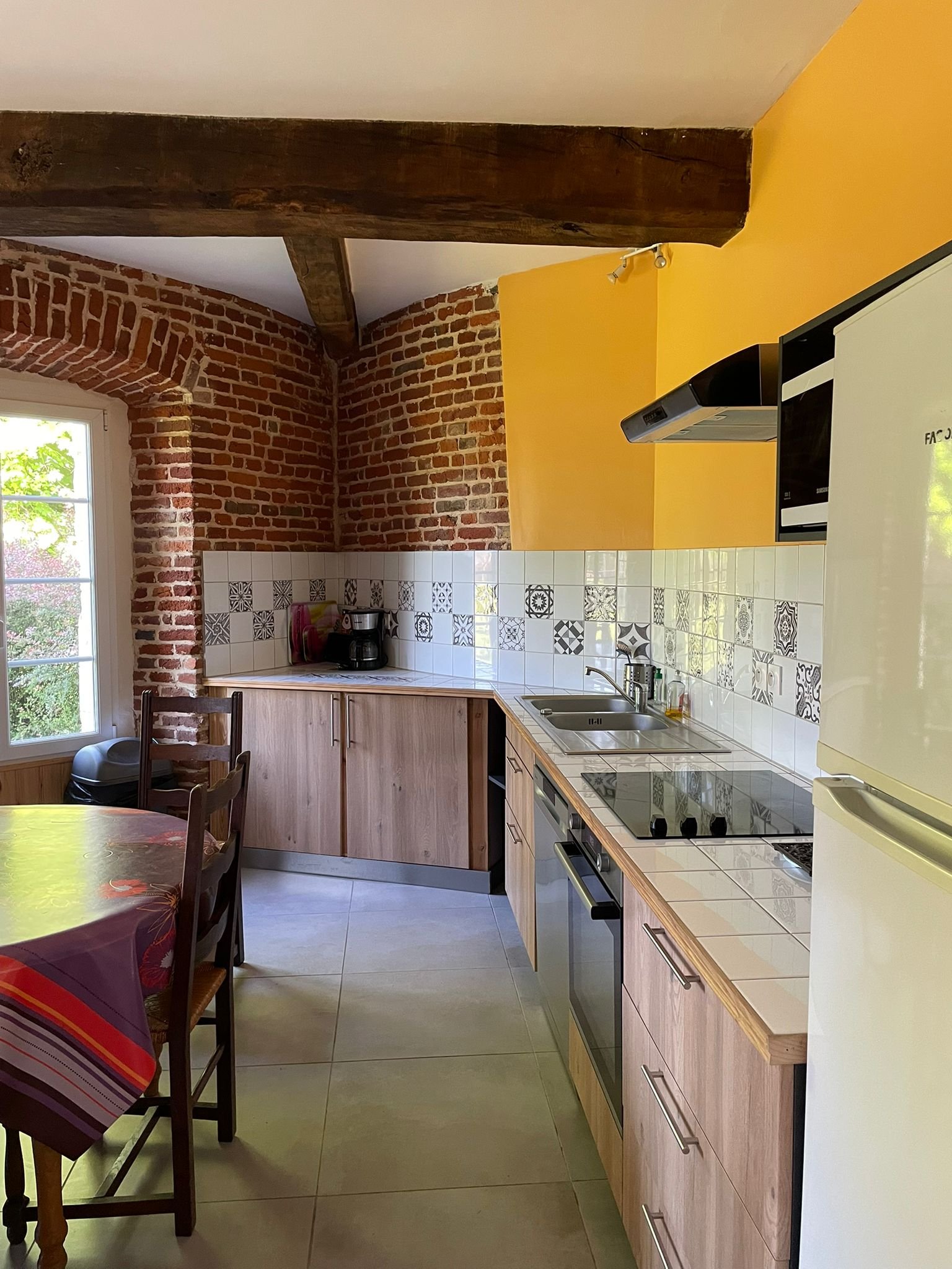 Cuisine avec murs en briques rouges, un plafond en bois, et un mur jaune. Combinaison de meubles en bois clair, évier, cuisinière électrique, et un réfrigérateur blanc. Une table avec porte-serviettes colorés et chaises en bois.