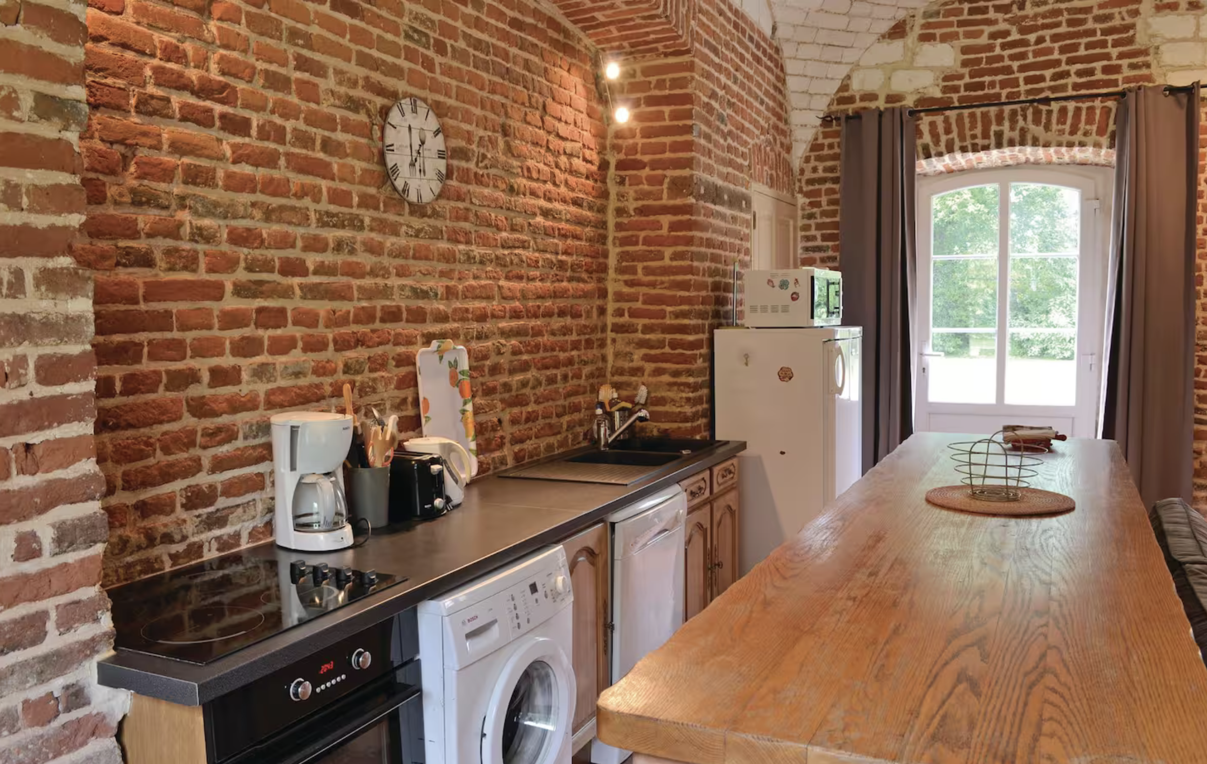 Cuisine avec un mur en briques rouges, un horloge, un comptoir en bois, un évier, un four, un lave-linge, un réfrigérateur, un micro-ondes, une fenêtre avec des rideaux gris, et une table en bois au centre.