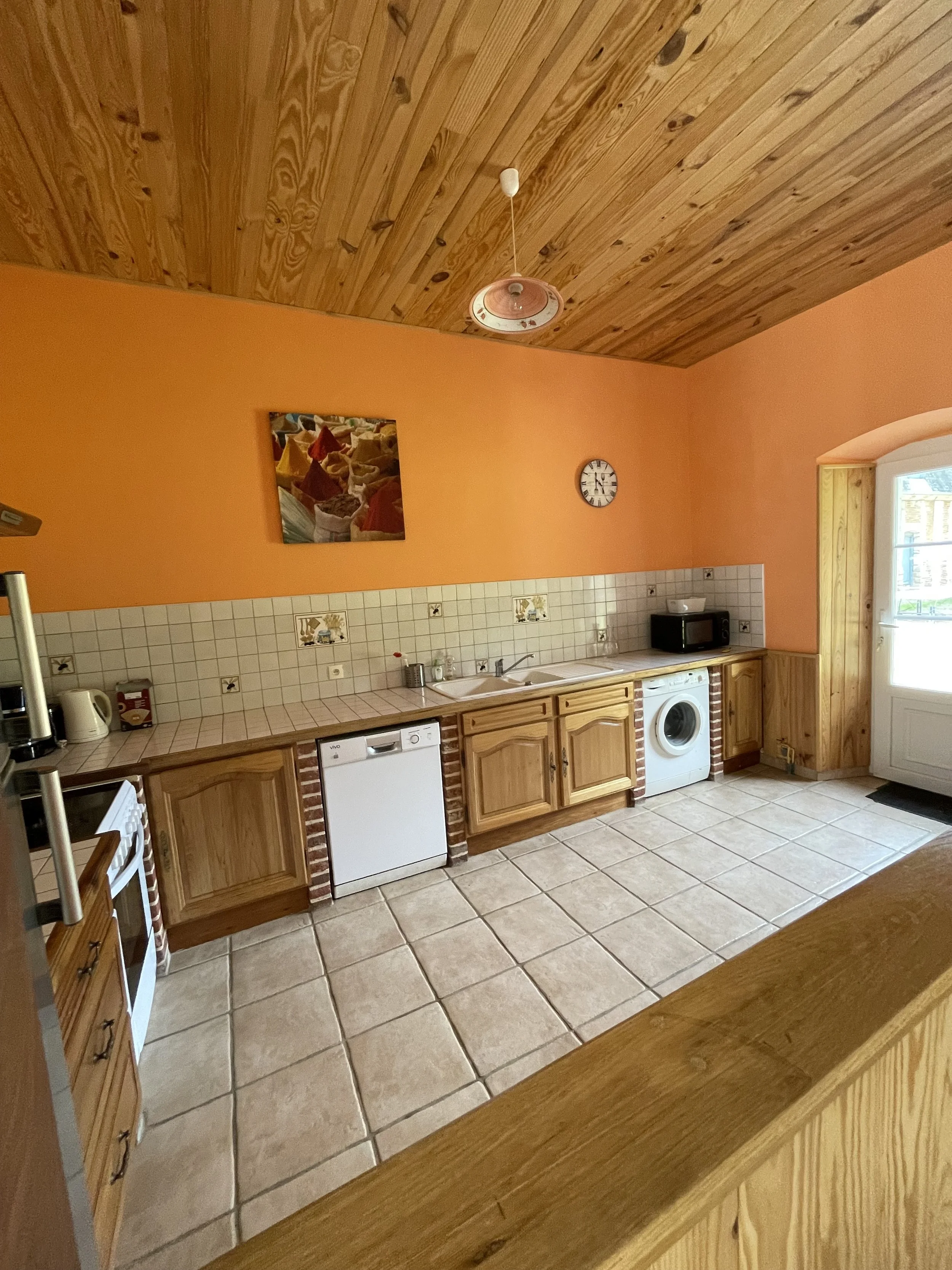 Cuisine avec murs en orange, plafond en bois, comptoir en bois, carrelage beige, machine à laver, micro-ondes, évier, plaque de cuisson, horloge
