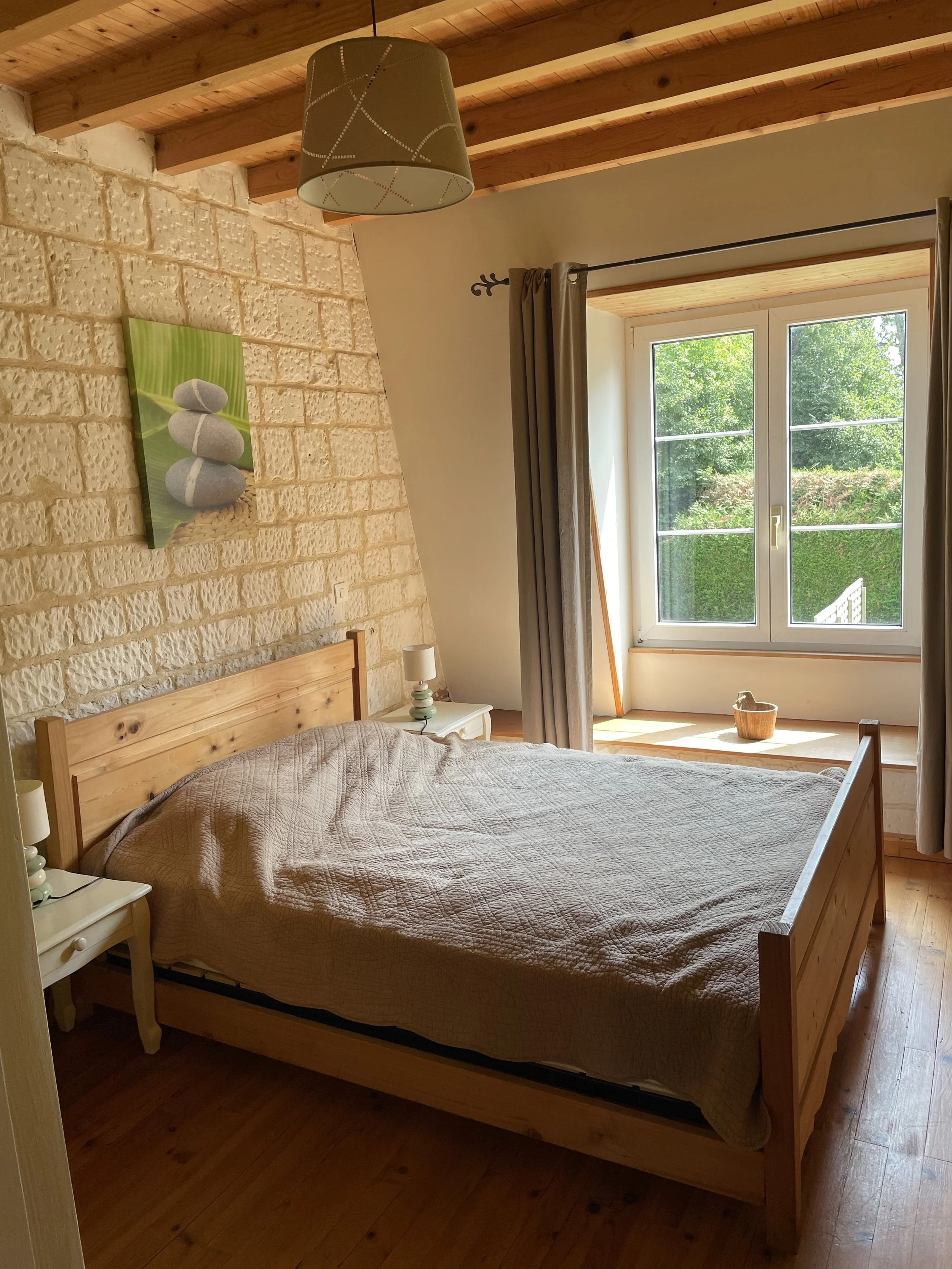 Chambre avec lit en bois, mur en pierre, tableau avec pierres empilées, grande fenêtre avec vue sur la verdure, lampes de chevet, lampadaire suspendu, sol en bois.