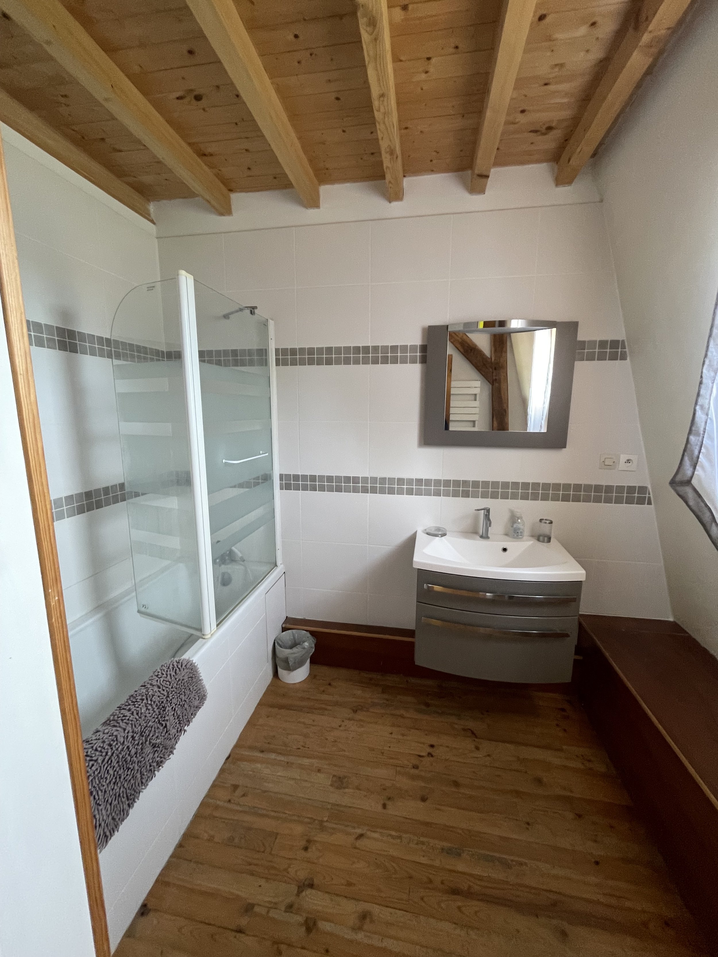 Une salle de bain avec un plafond en bois, un mur avec des carreaux blancs et une bande de carreaux gris, un lavabo avec un miroir, une cabine de douche avec parois en verre, un tapis gris, un petit poubelle, et un banc en bois.