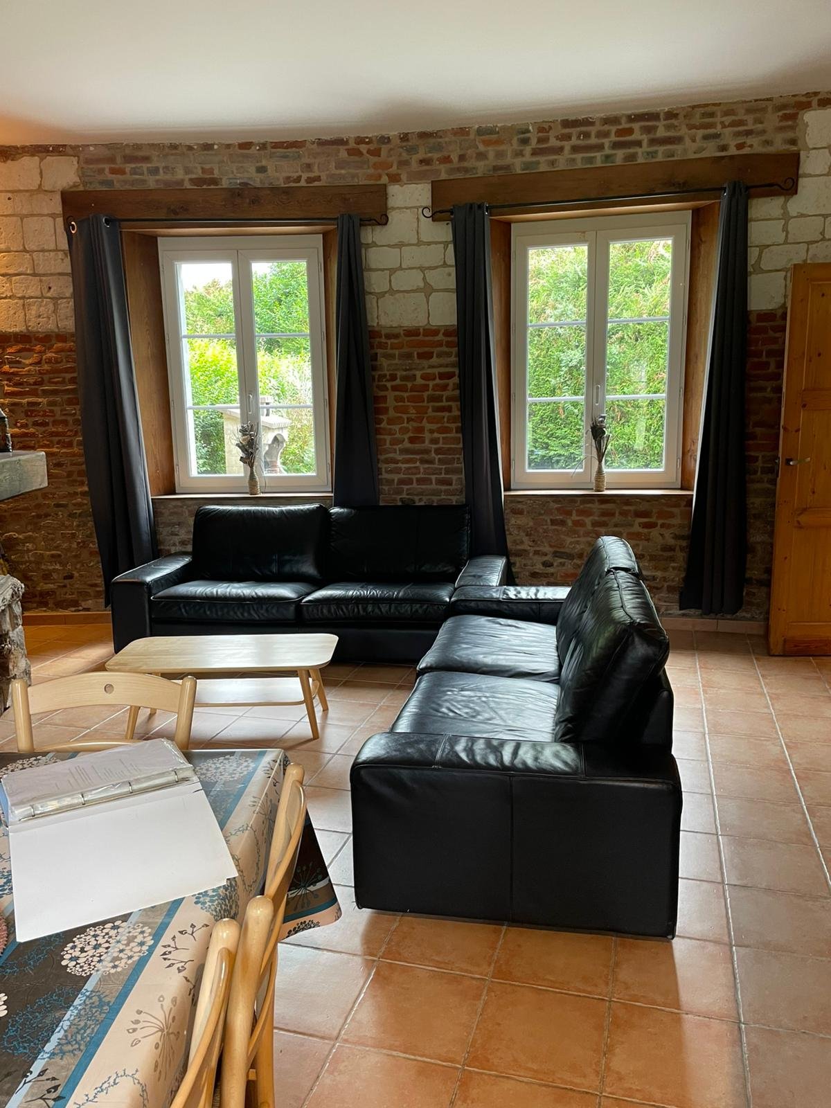 Salon avec deux canapés en cuir noir, deux fenêtres avec volets en bois et rideaux noirs, murs en briques, table basse en bois claire, mobilier en bois naturel, table avec un livre ouvert et des chaises en bois