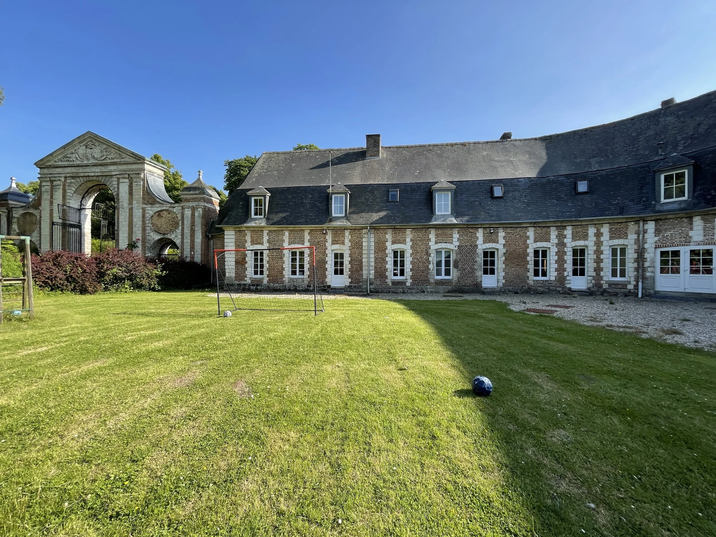 Vue d'une maison en pierre avec un grand jardin verdoyant, un but de football, et un ballon sur l'herbe. Le ciel est clair et bleu.