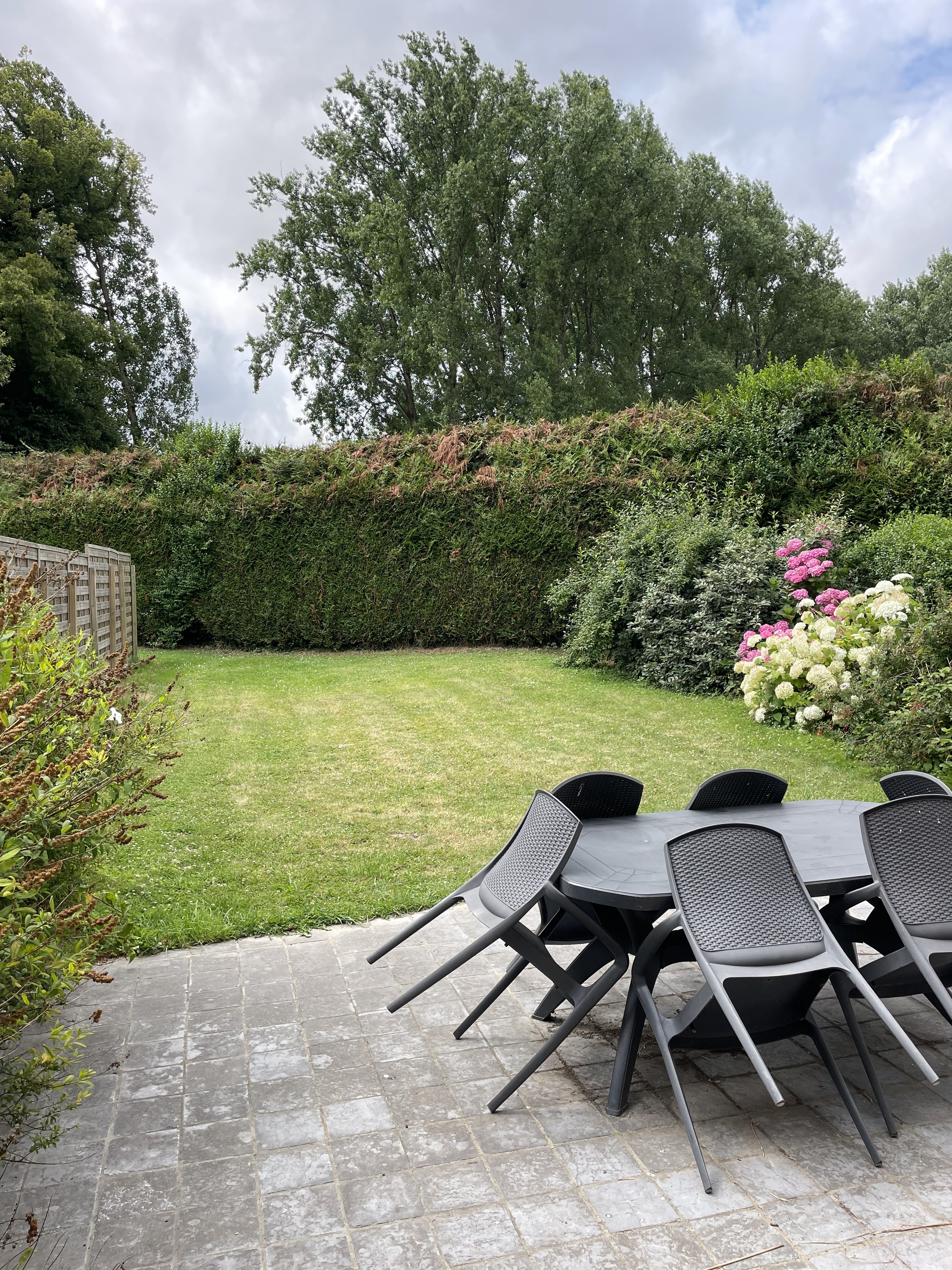Jardinière avec une table en plastique entourée de plusieurs chaises en plastique noir, sur une terrasse pavée, avec un jardin bien entretenu arboré et des buissons fleuris, ciel nuageux.