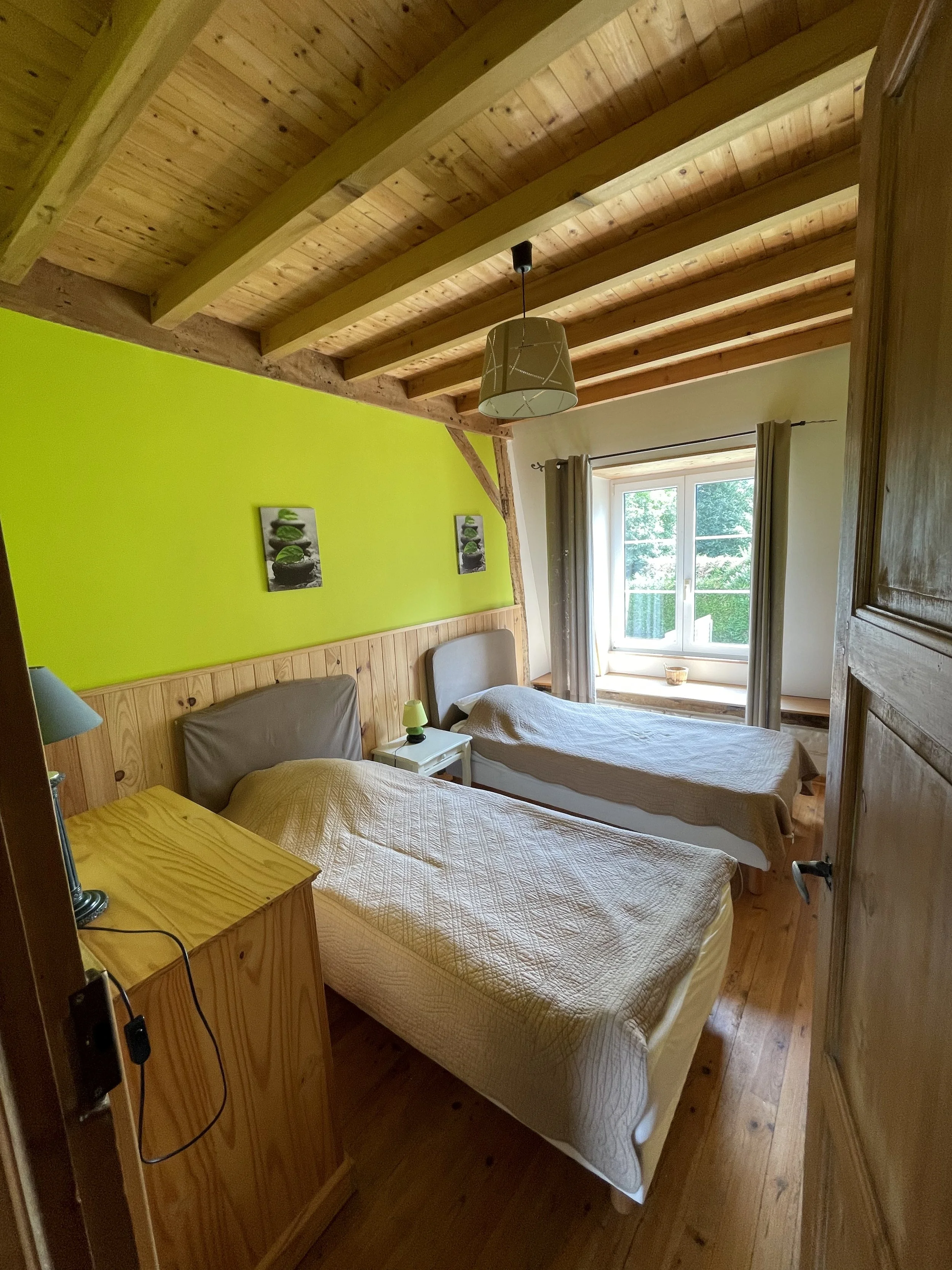 Chambre avec deux lits simples, mur vert, fenêtres avec vue sur la nature, mobilier en bois.