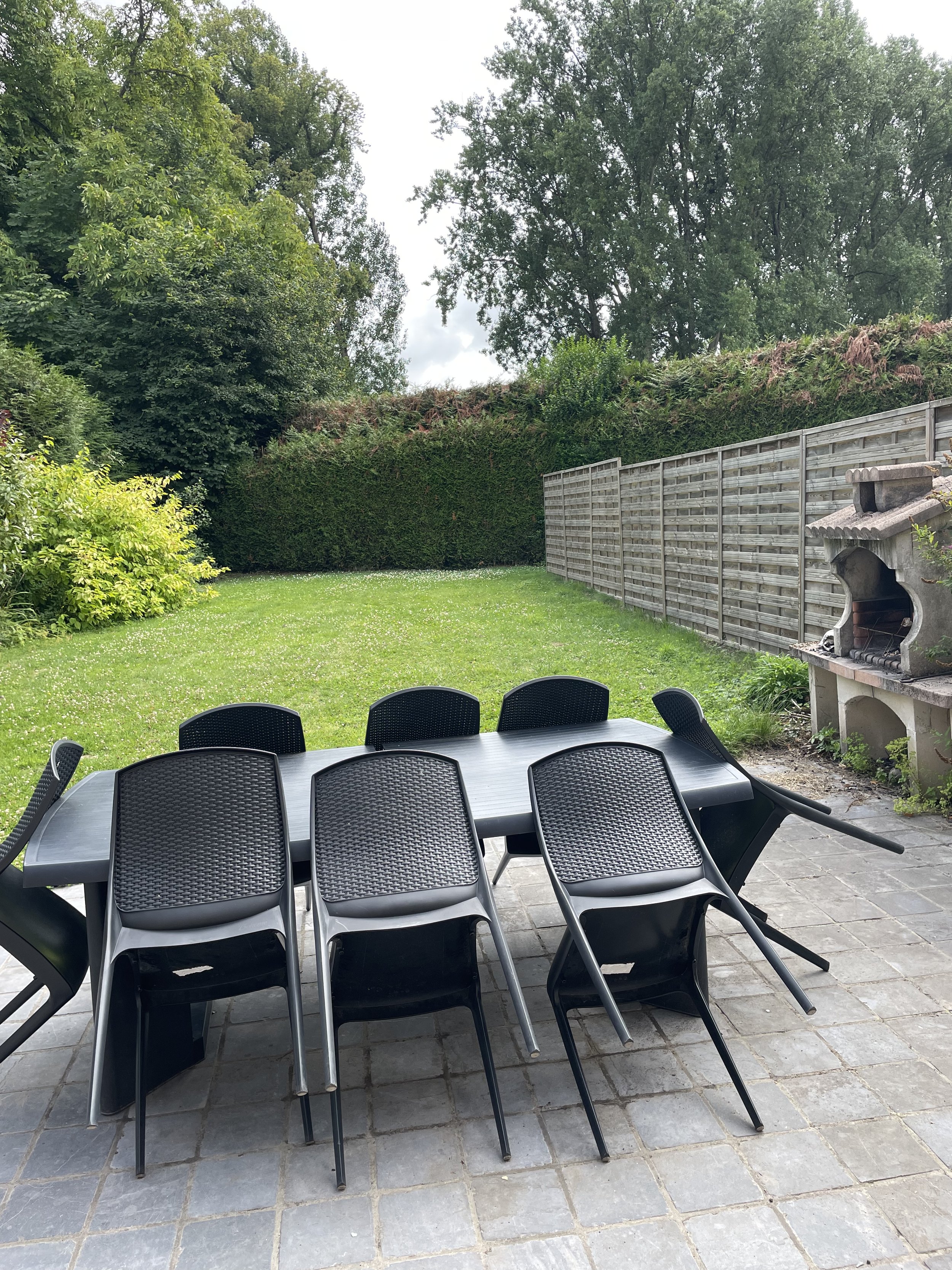 Table en plastique noir avec huit chaises noires en plastique autour, situé sur une terrasse pavée en pierre, avec un jardin herbeux en arrière-plan et une cheminée en pierre sur le côté droit.