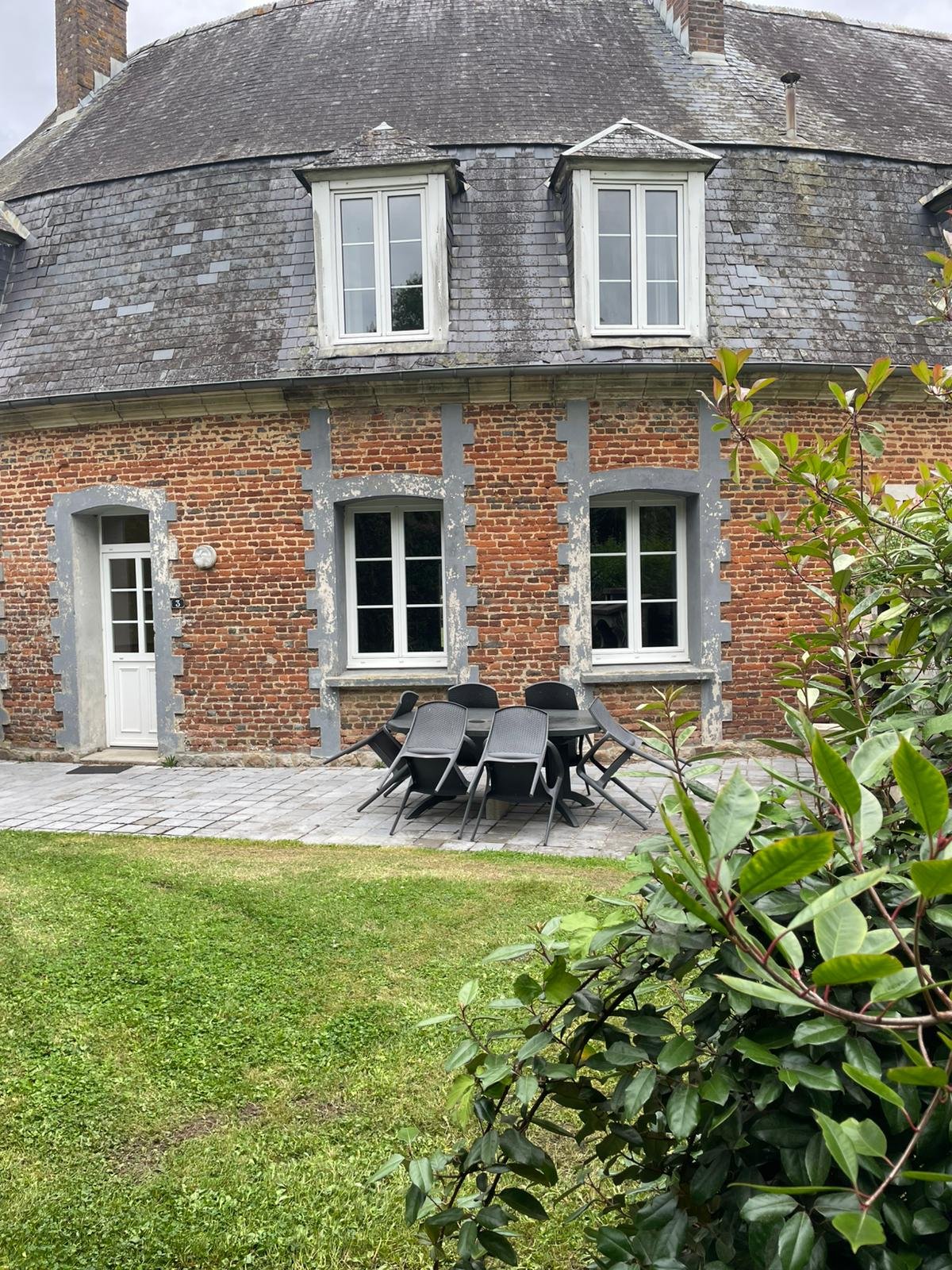 Une maison en briques avec des fenêtres blanches et des volets, au rez-de-chaussée une terrasse avec une table et des chaises en plastique noir, un mur en pierre et un toit en ardoise, devant un jardin avec de l'herbe et des arbustes.