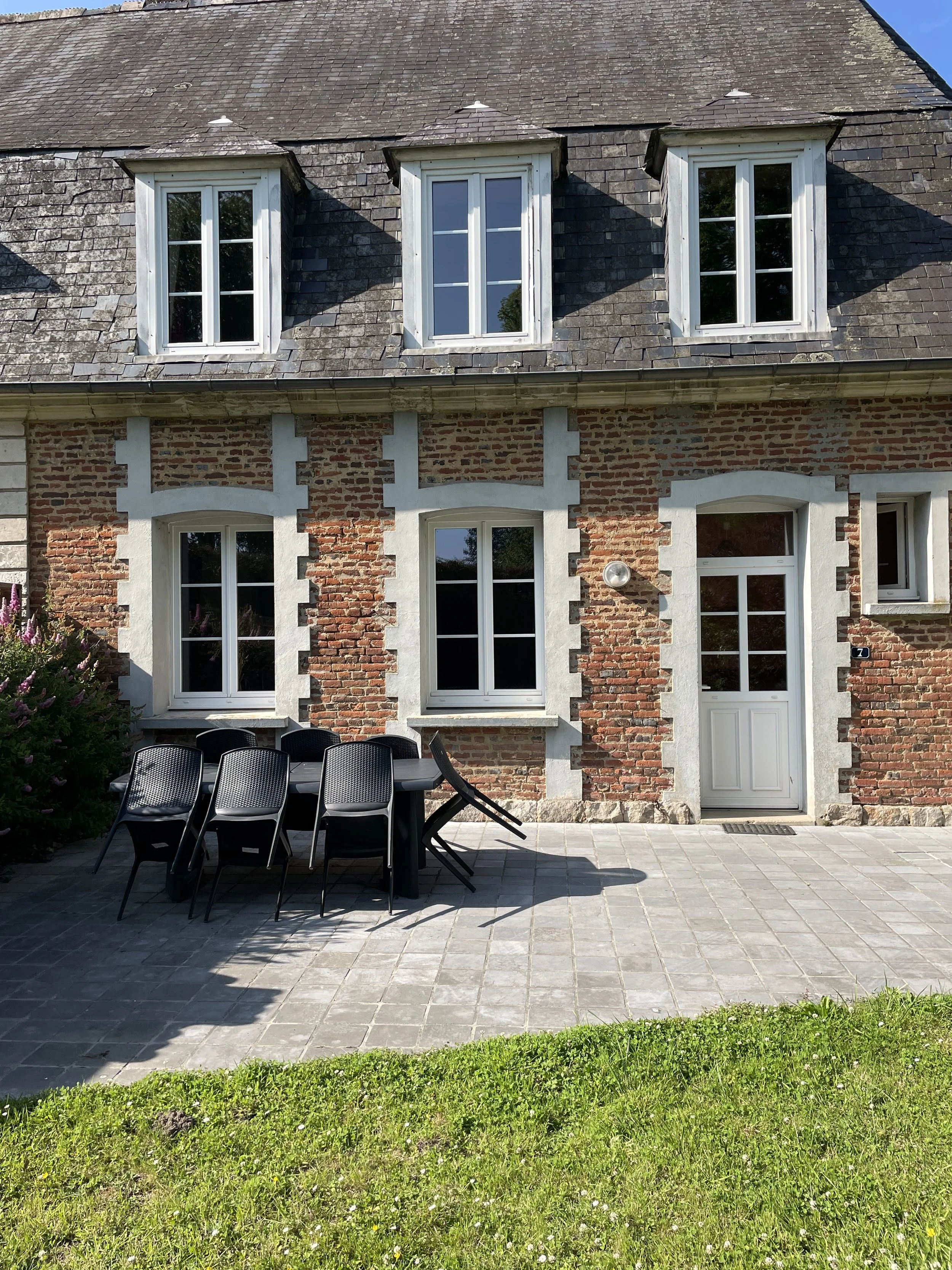 Une maison en briques avec des fenêtres blanches et un toit en ardoise. Un patio avec une table et des chaises noires au premier plan. Il y a un peu d'herbe et des plantes d'arbustes en coin.
