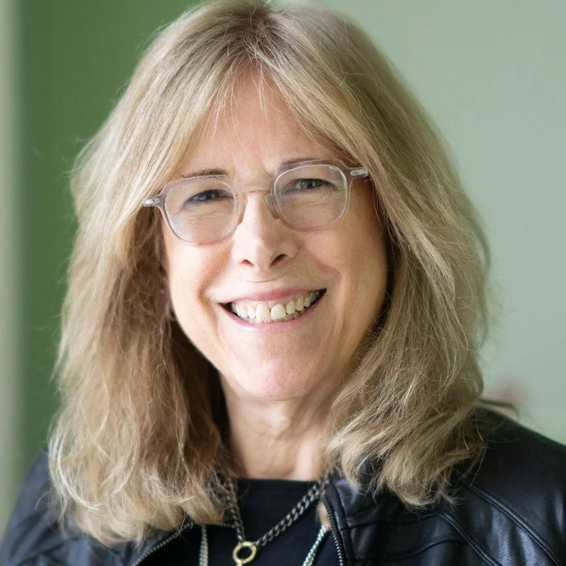 Cheryl Kiser, Strategic Innovation Advisor: Portrait of a smiling woman with long blonde hair, wearing glasses and a black leather jacket, standing against a green background.