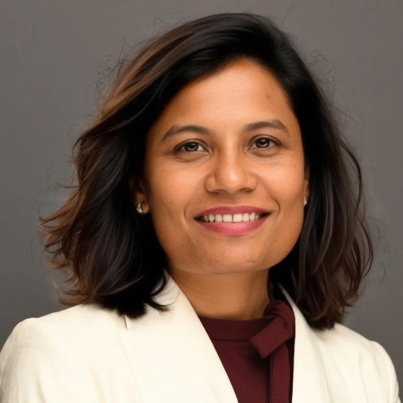 Smitha Gudapakkam, Chief Executive Offer: A woman with shoulder-length dark hair, wearing a white blazer and maroon top, smiling against a gray background.