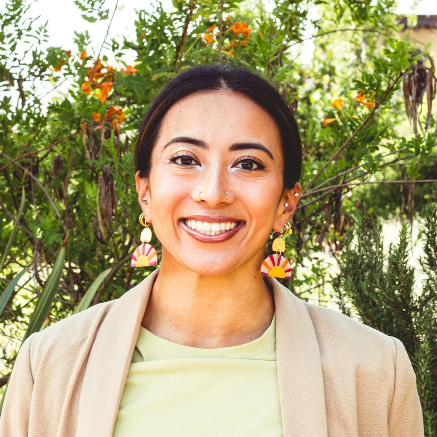 Rima Sheehab, Integrative Health Coach and Strategic Product Lead: A smiling woman with dark hair, wearing a green and white tropical print blouse, standing outdoors in front of a backdrop of tall green plants.