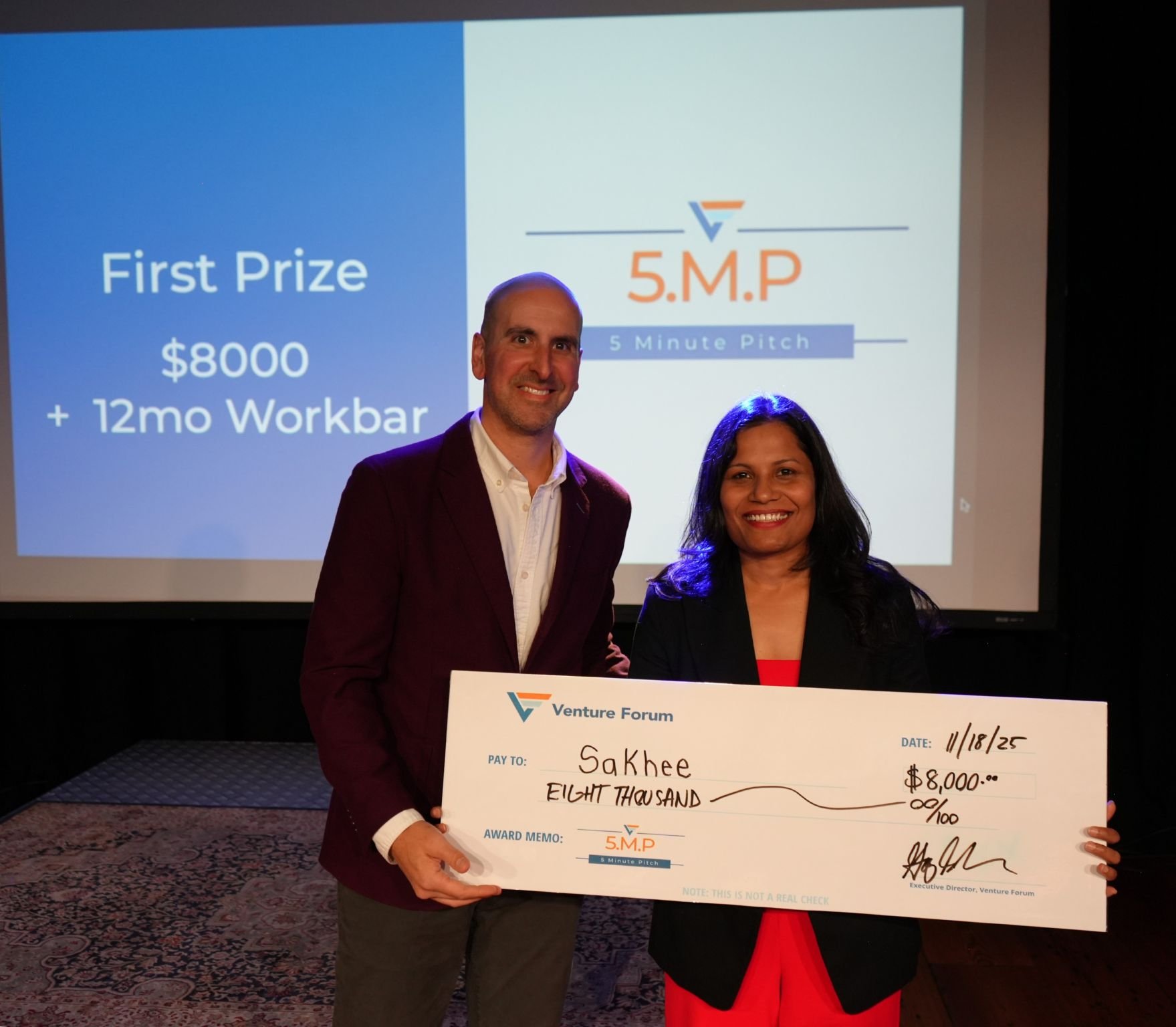  Smitha Gudapakkam, founder of Sakhee, stands onstage at the Venture Forum 5‑Minute Pitch Competition holding a check for $8,000 in first‑place funding, with the event screen behind her displaying “First Prize” and the 5‑Minute Pitch logo.
