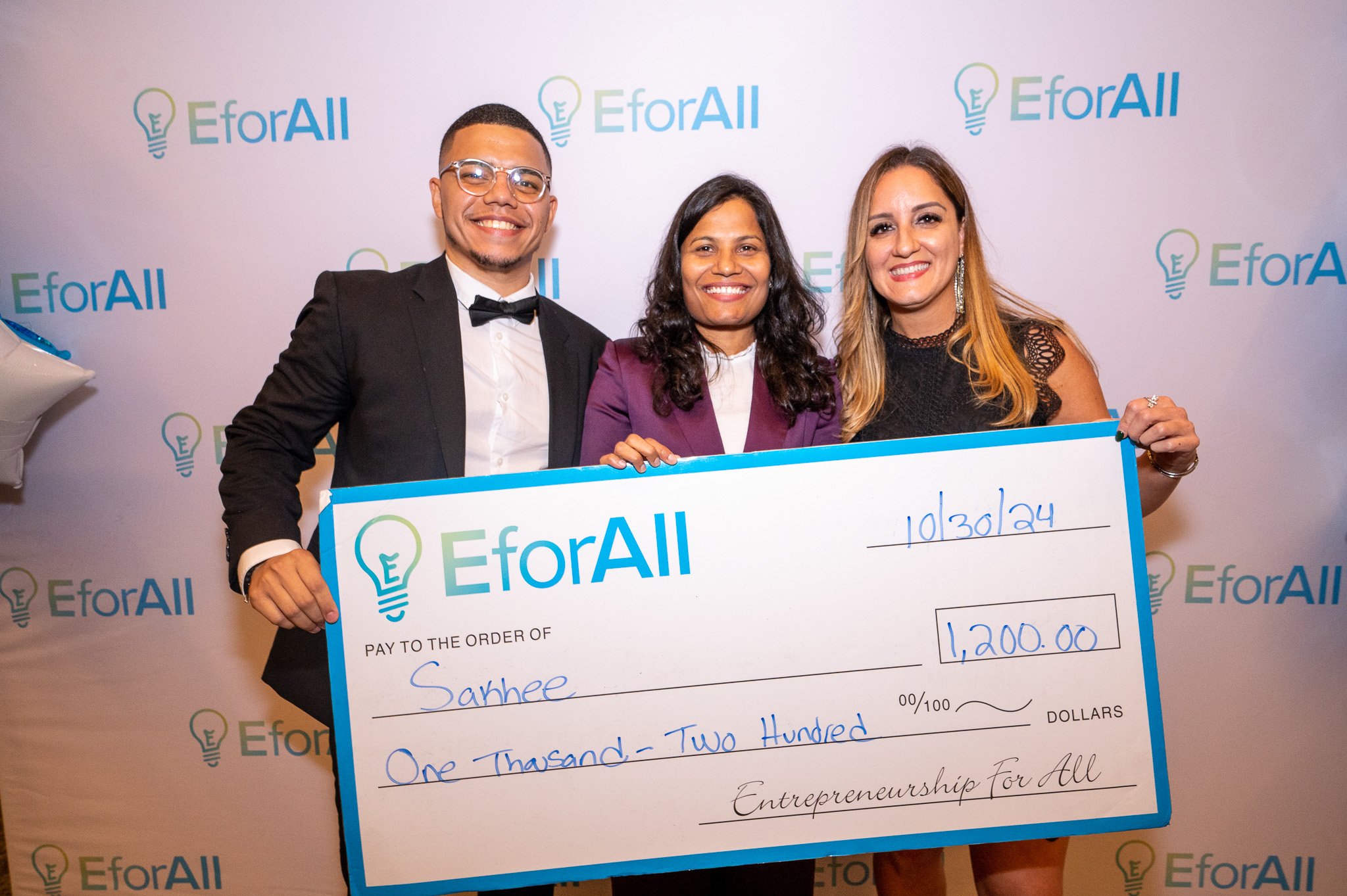 Three people in formal attire holding a large ceremonial check for $1,200.00 made out to Sakhee, with a backdrop displaying EforAll logos. Smitha Gudapakkam is in the middle.