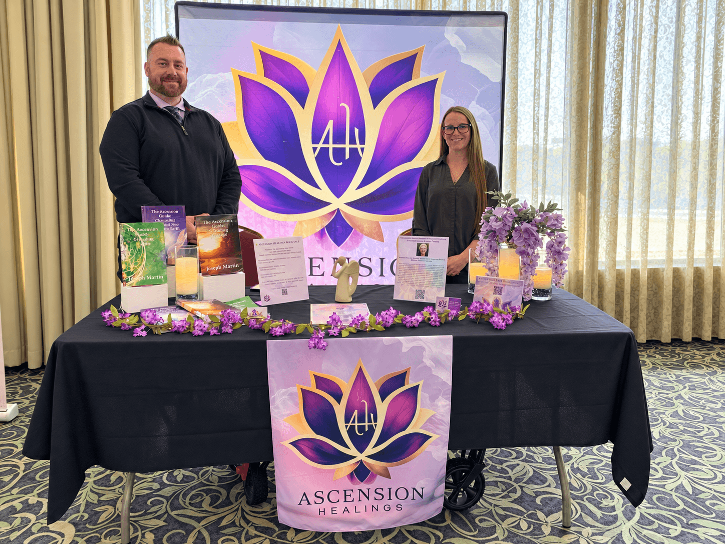 Joseph and Jennifer Martin standing behind an Ascension Healings booth with purple lotus branding, books, candles, flowers, and informational displays.