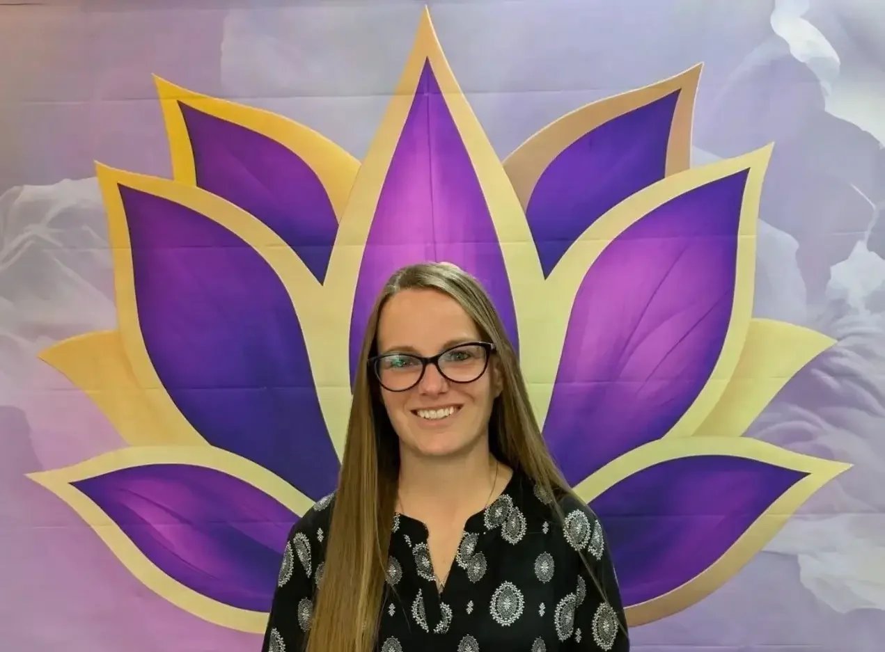 Jennifer Martin co founder and Reiki master with long blonde hair, wearing glasses and a black blouse with white patterns, standing in front of a large purple and yellow lotus-shaped backdrop.