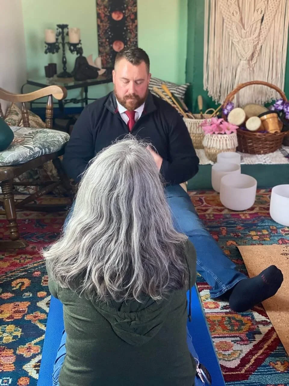 Joseph Martin qhht practitioner and author and a woman client with gray hair sitting on a blue yoga mat in a room decorated with colorful rugs, baskets, and candles, engaging in a healing or therapy session.