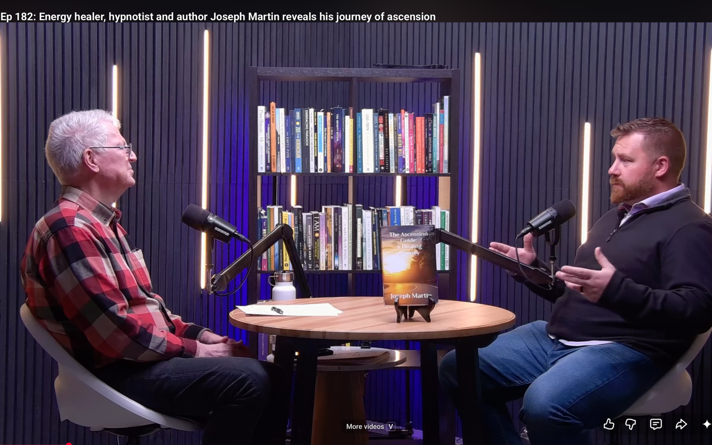 Two men sitting across from each other engaged in a conversation with microphones on stands. A wooden table with a book titled 'The Ascension Guide: A Healing Journey' by Joseph Martin, a white water bottle, and sheets of paper are on the table. Behind them is a bookshelf filled with various books, illuminated by vertical lights on a dark, textured wall. The headline above reads, 'Ep 182: Energy healer, hypnotist and author Joseph Martin reveals his journey of ascension.'