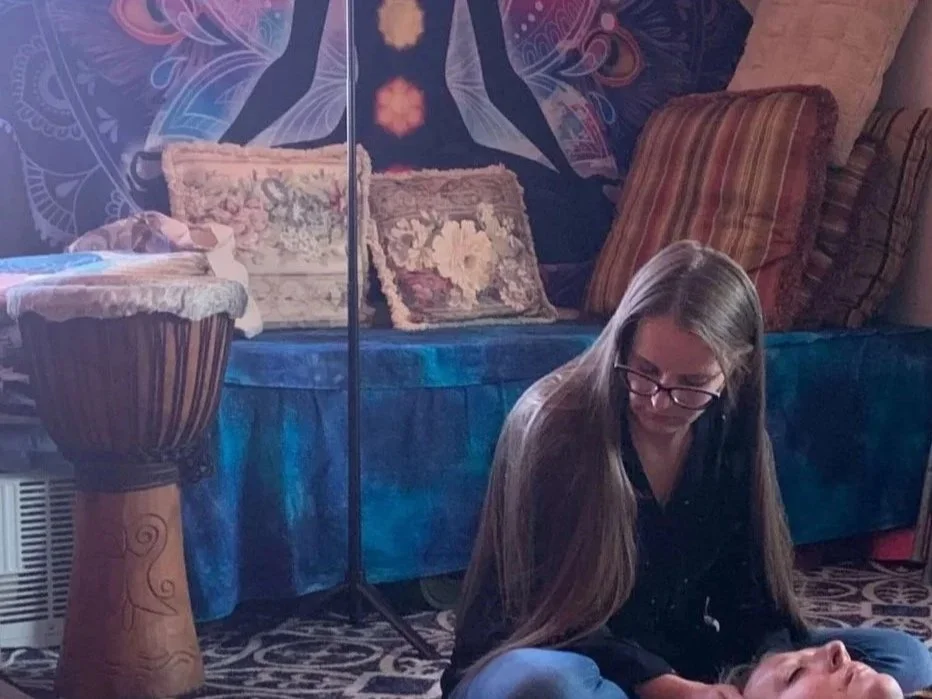 Jennifer Martin reiki master is sitting on the floor next to a person lying down. The background features cushions and a tapestry with a fantasy and celestial design, including an angelic figure and glowing symbols.