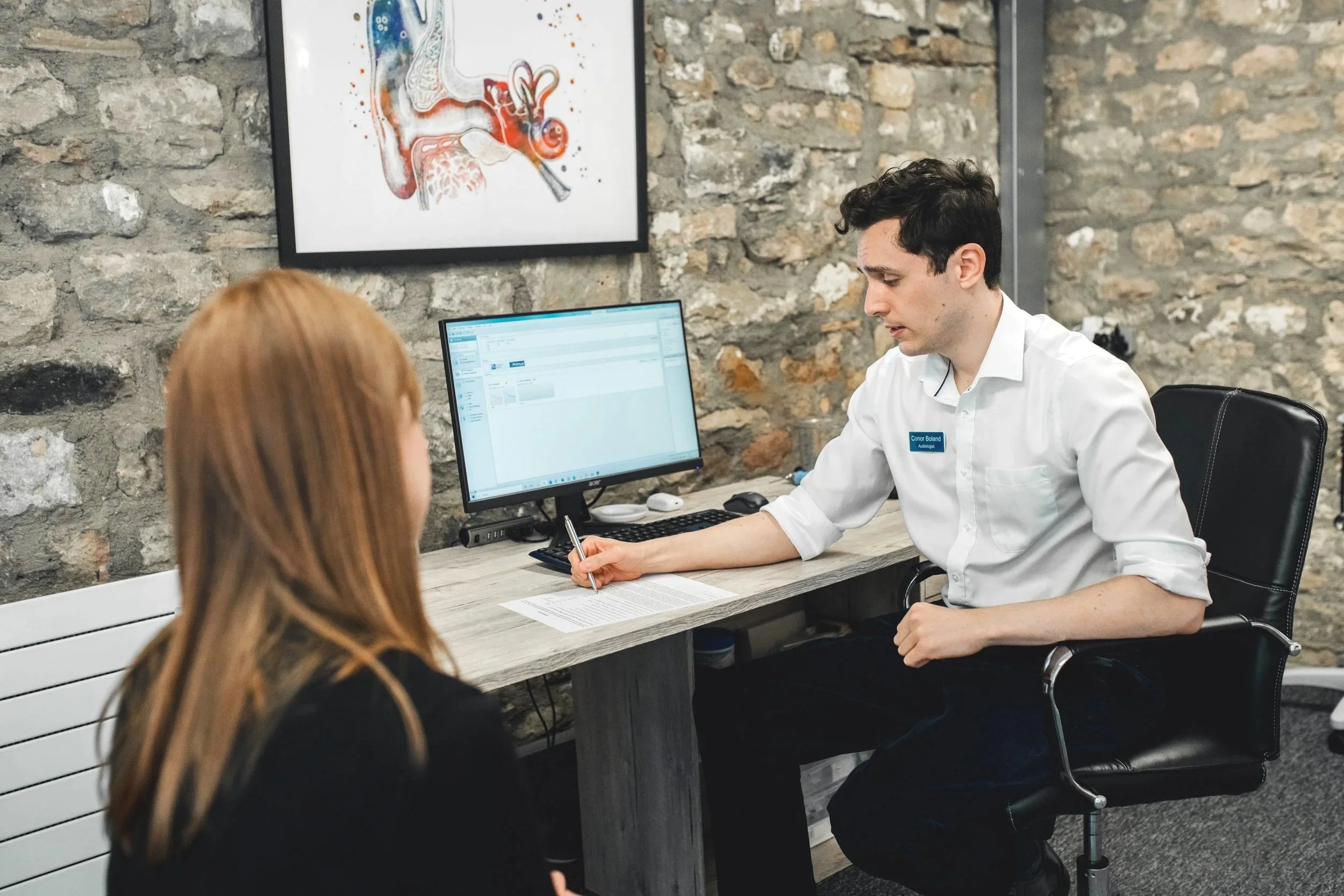 A veterinary consultation with a male veterinarian at a desk, using a computer, talking to a woman with red hair in a black shirt. The veterinarian is wearing a white coat with a name tag. The background has a stone wall and a framed anatomical diagram of the ear.