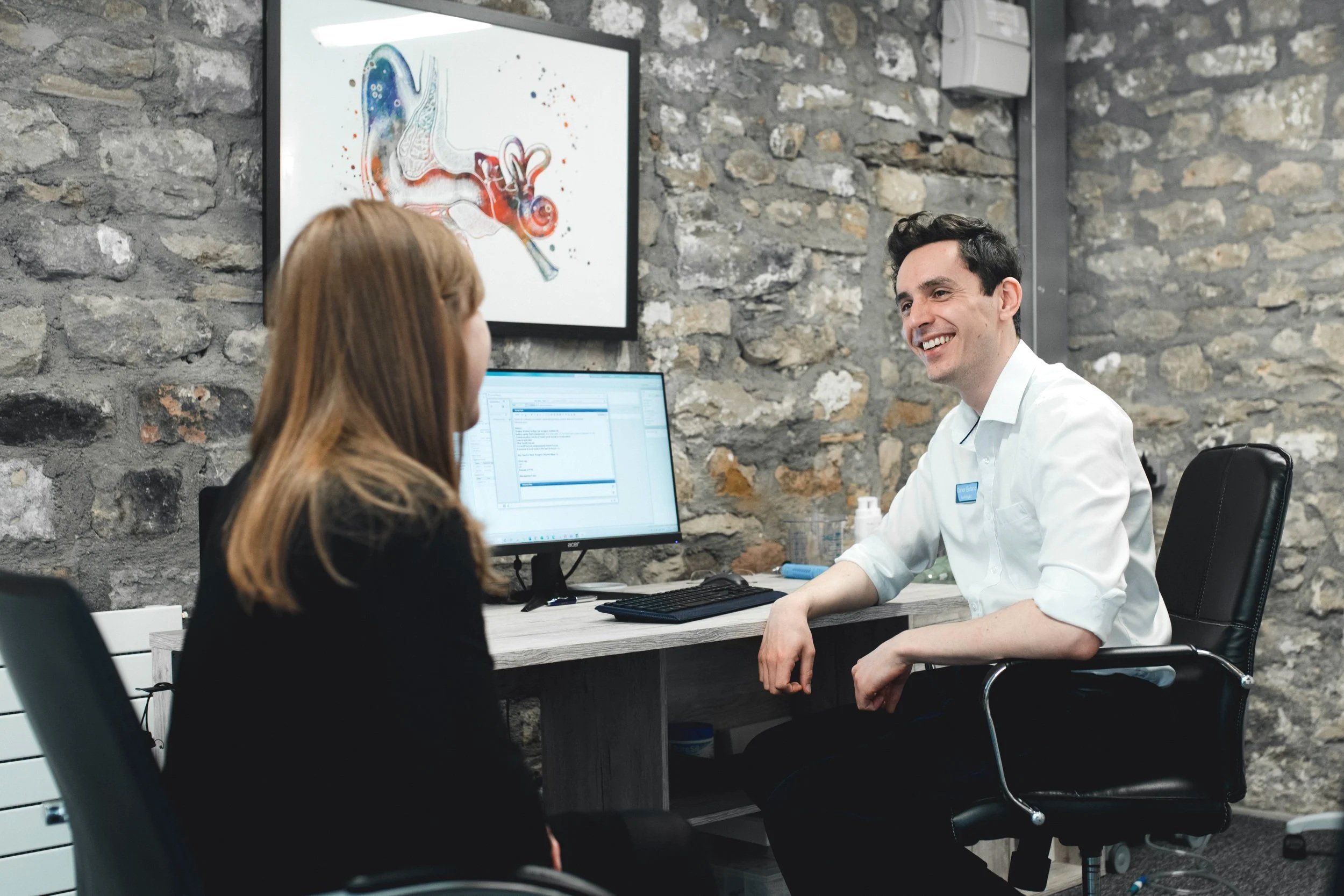 A medical consultation taking place in a clinic with a stone wall background, featuring a female patient with long reddish-brown hair and a male doctor with dark hair in a white shirt, smiling at each other across a desk with computer monitors.