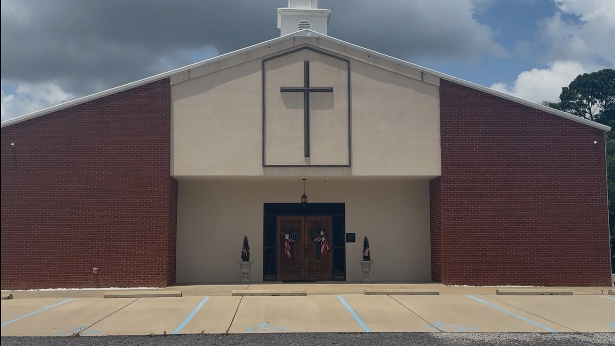 Front entrance of a church with a large cross above the door, decorated with patriotic wreaths and American flags, and an empty parking lot in the foreground.