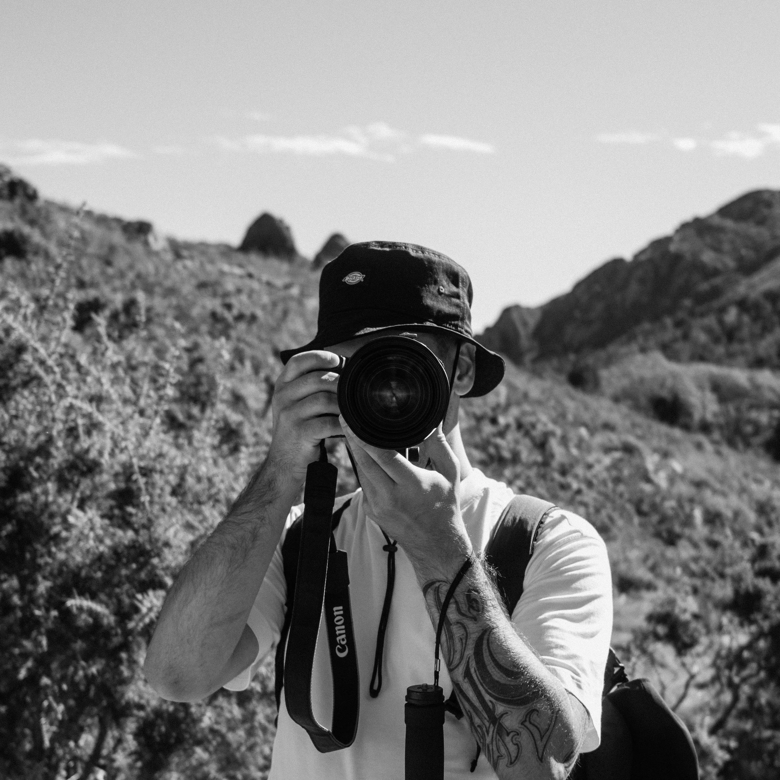 Une personne en plein air, portant un chapeau et un t-shirt blanc, se prend en photo avec un appareil photo reflex, avec un paysage de montagnes et de végétation en arrière-plan.
