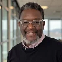 A middle-aged man with glasses and a beard, smiling, in a professional setting with glass windows in the background.