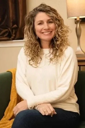 A woman with curly blonde hair smiling, sitting in a cozy room with a lamp and dark curtains in the background.