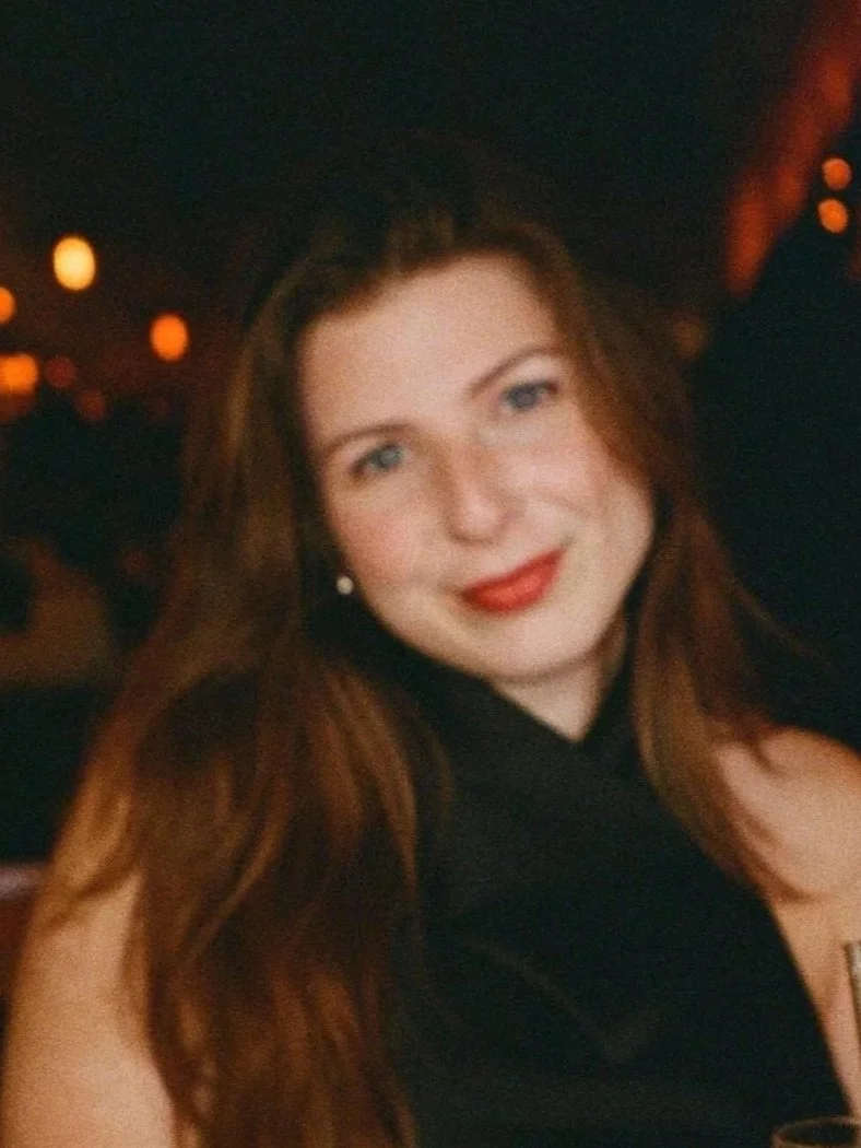 A woman with long, wavy red hair and fair skin smiling at the camera, wearing a black top, with a dimly lit background and orange bokeh lights.