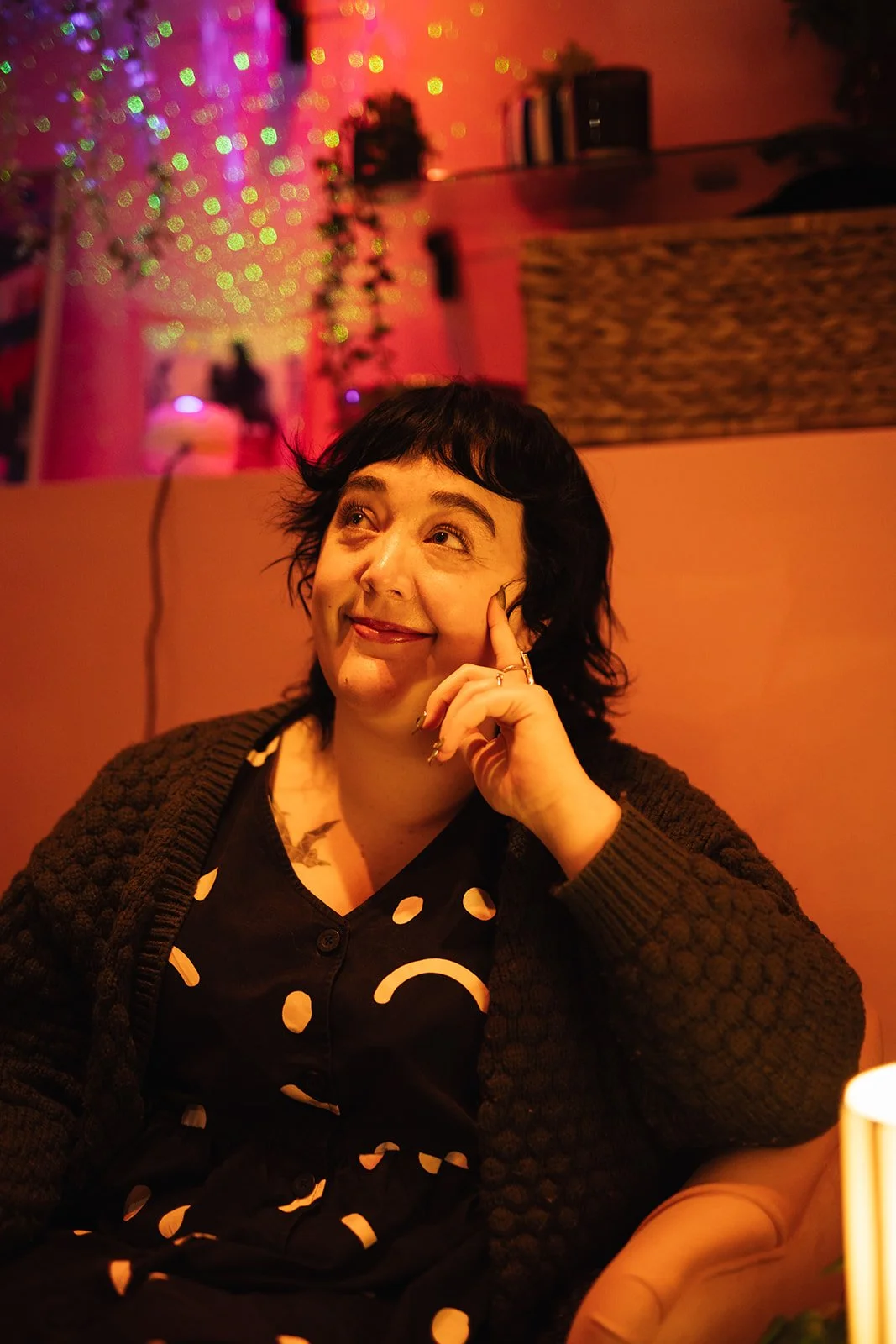 a white woman with dark hair looks into the distance, she is sat in a pink counselling room