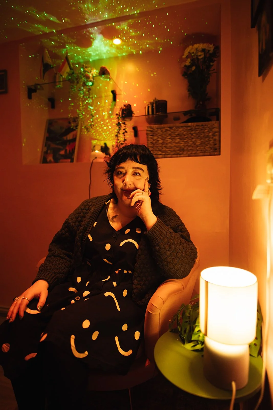 a white woman wearing a patterned black dress and green cardigan. She looks into the camera and smiles.