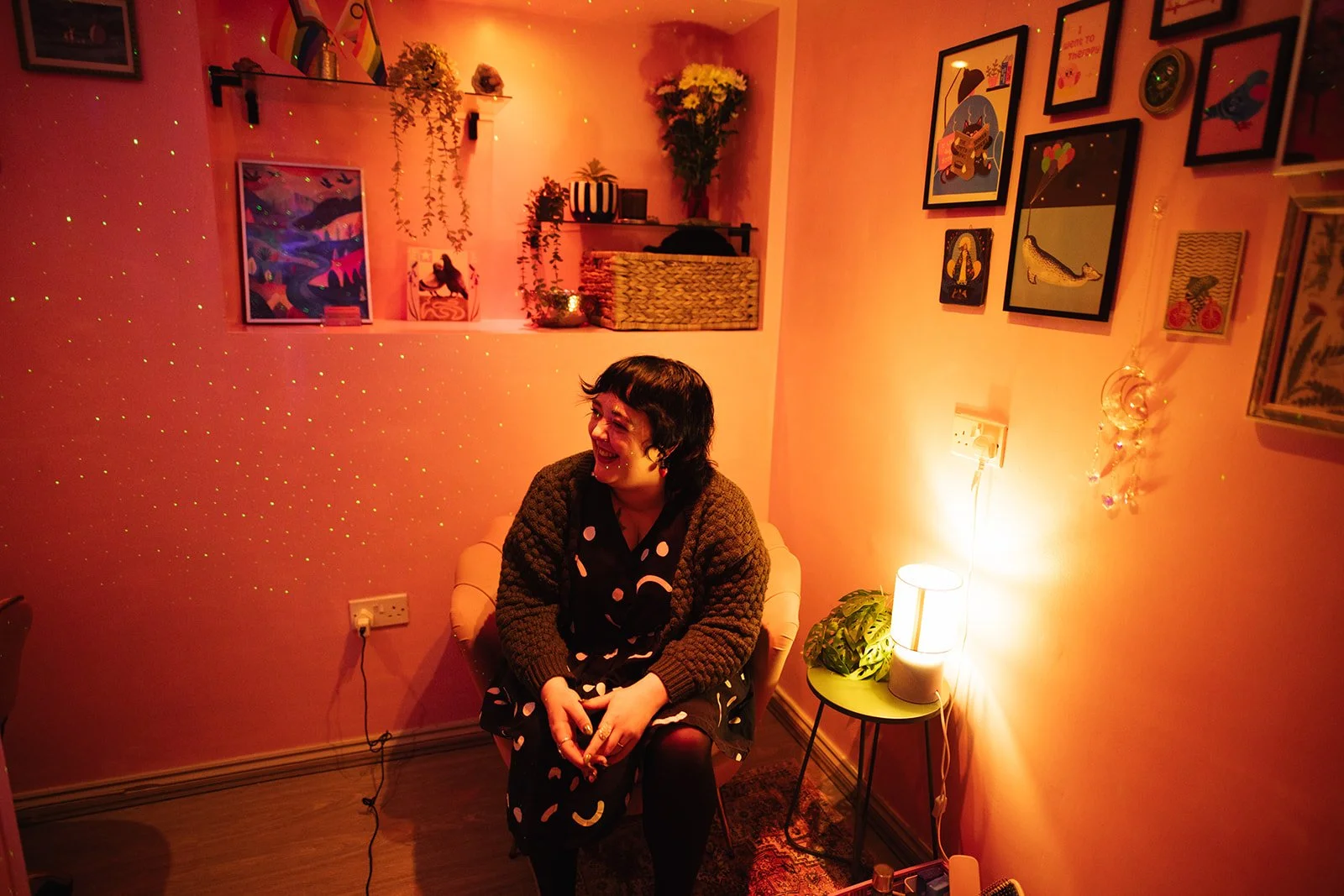 a white woman with curly dark hair is seated in a pink armchair, in a pink room. She is laughing as she looks into the distance.