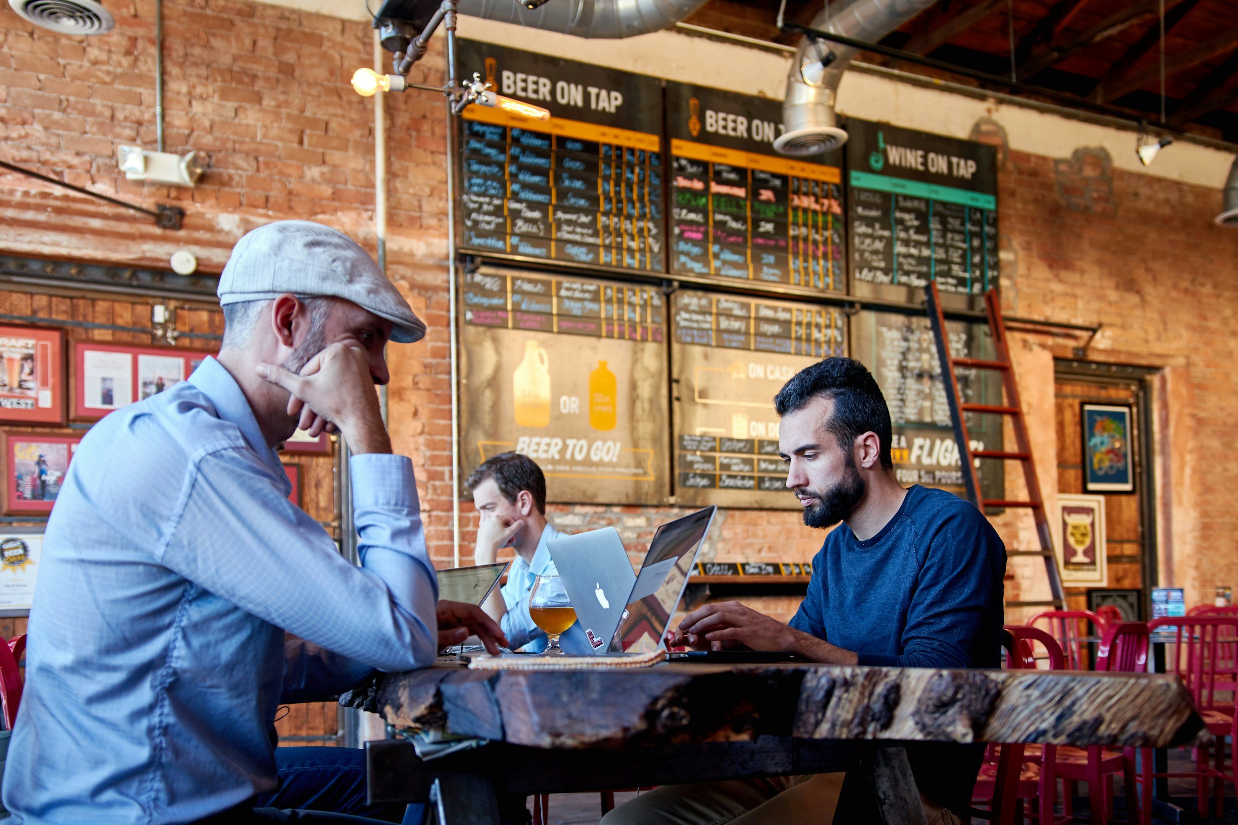 Three men sitting at a bar-height table in a restaurant with a brick wall background, looking at their laptops. The man on the left, wearing a light gray hat and blue shirt, has his hand on his face. The man on the right, with dark hair and a beard, is focused on his laptop. The restaurant has a rustic decor with framed pictures, a ladder, and a menu board on the wall.
