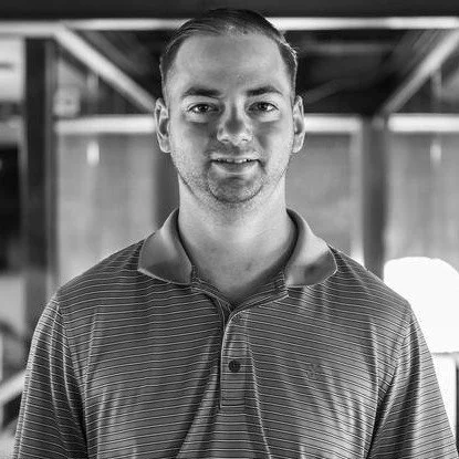 Black and white photo of a man wearing a polo shirt, smiling at the camera, indoor setting with structural beams in the background.