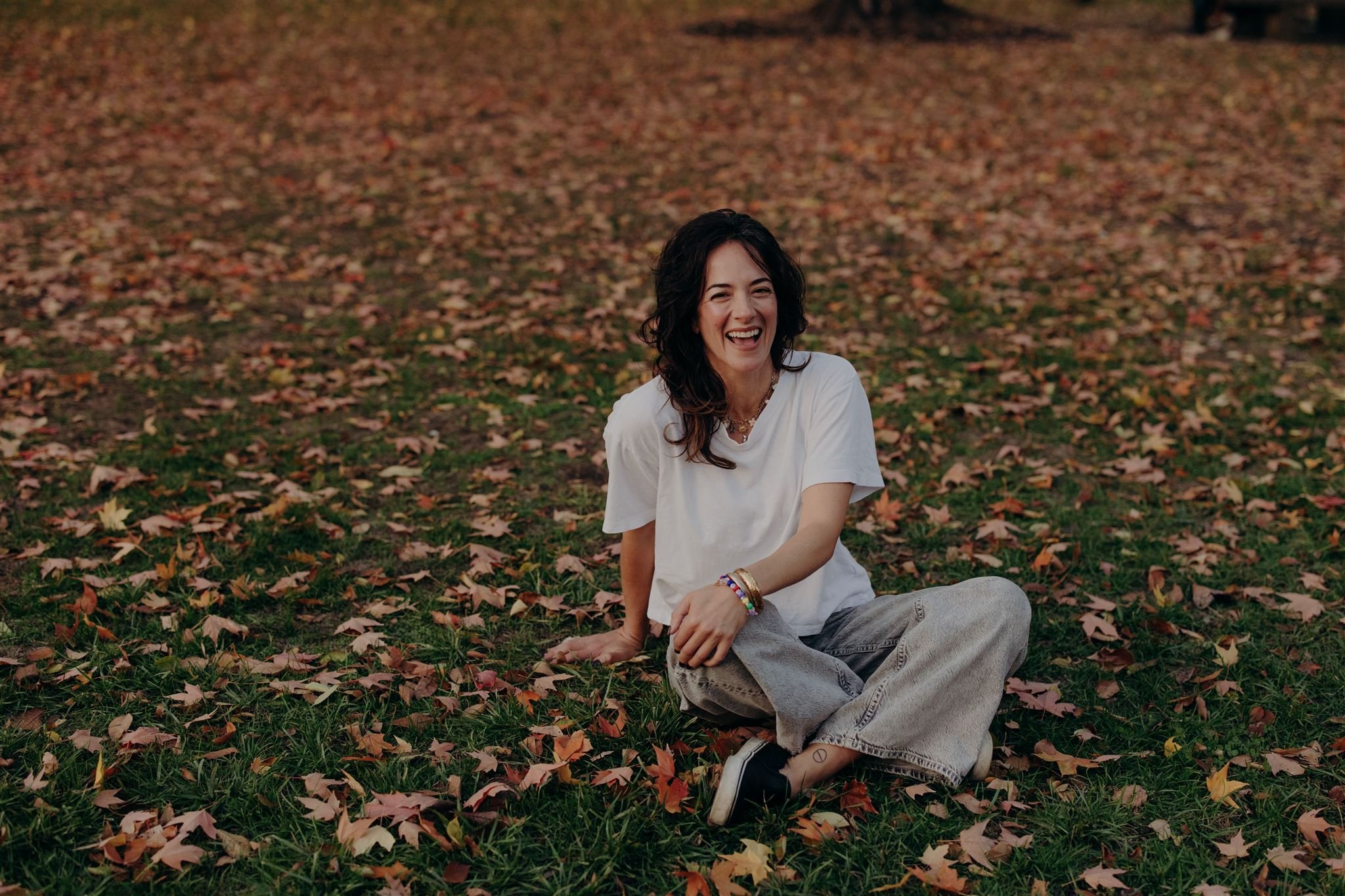 A woman with dark curly hair sitting on a grassy field covered with fallen autumn leaves, smiling and laughing.